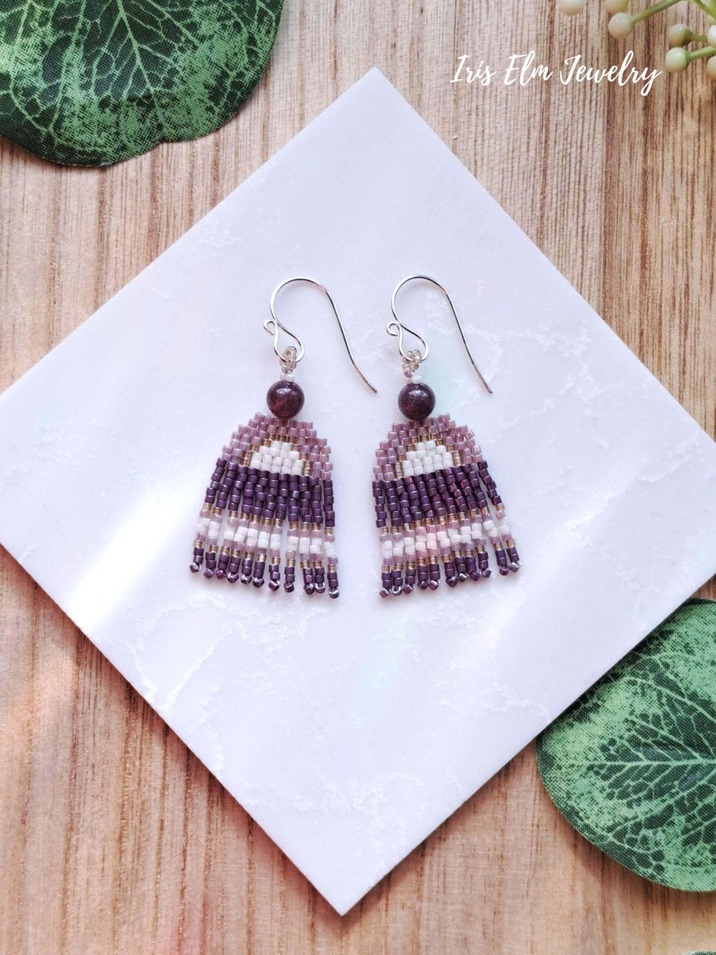 Pair of beaded lepidolite white and purple fringe seed bead earrings on a white card with 'Iris Elm Jewelry' branding, placed on a wooden surface with green leaves.