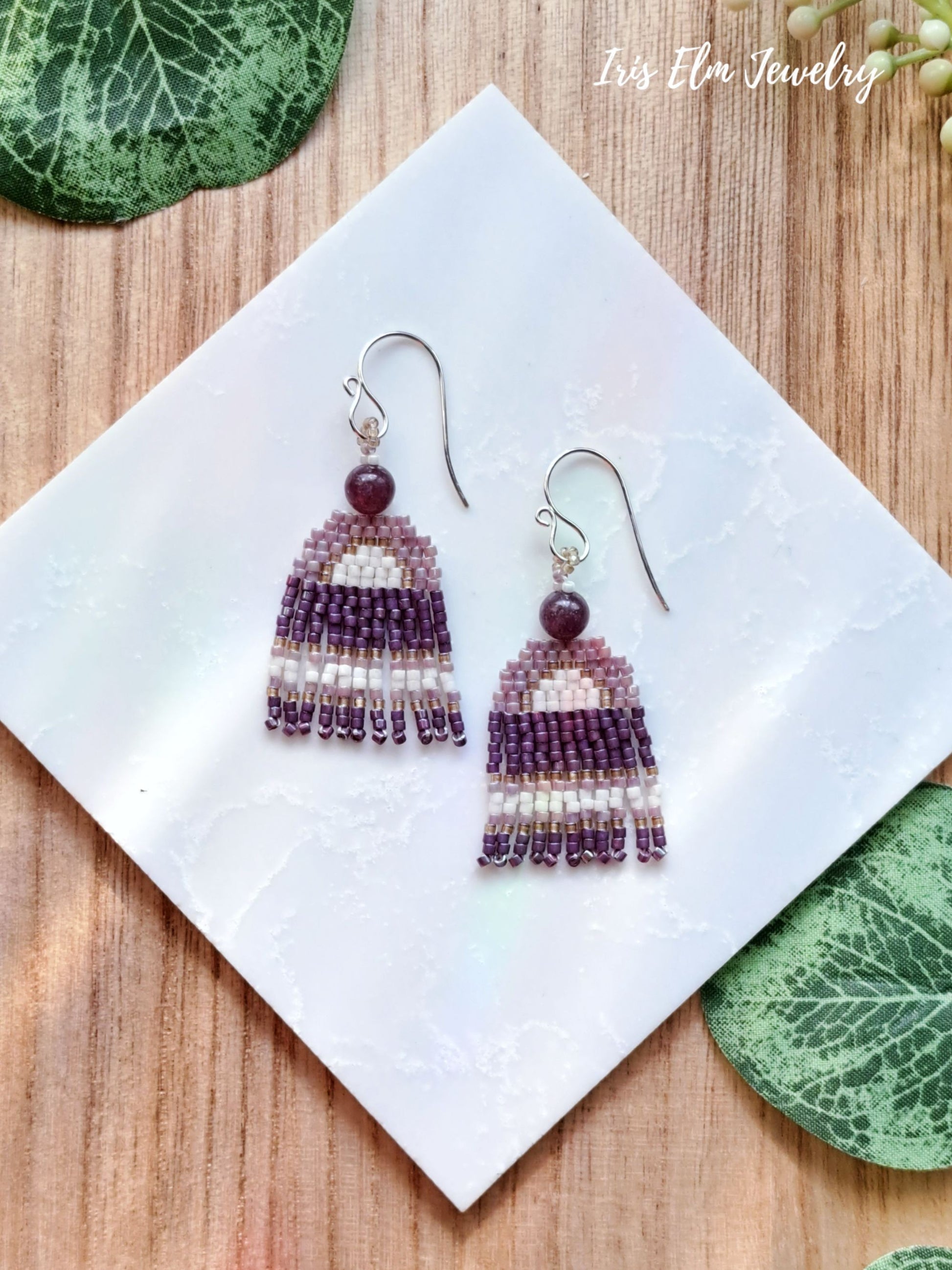 Beaded small seed bead fringe lepidolite earrings on a white square with green leaves on a wooden surface, featuring 'Iris Elm Jewelry'.