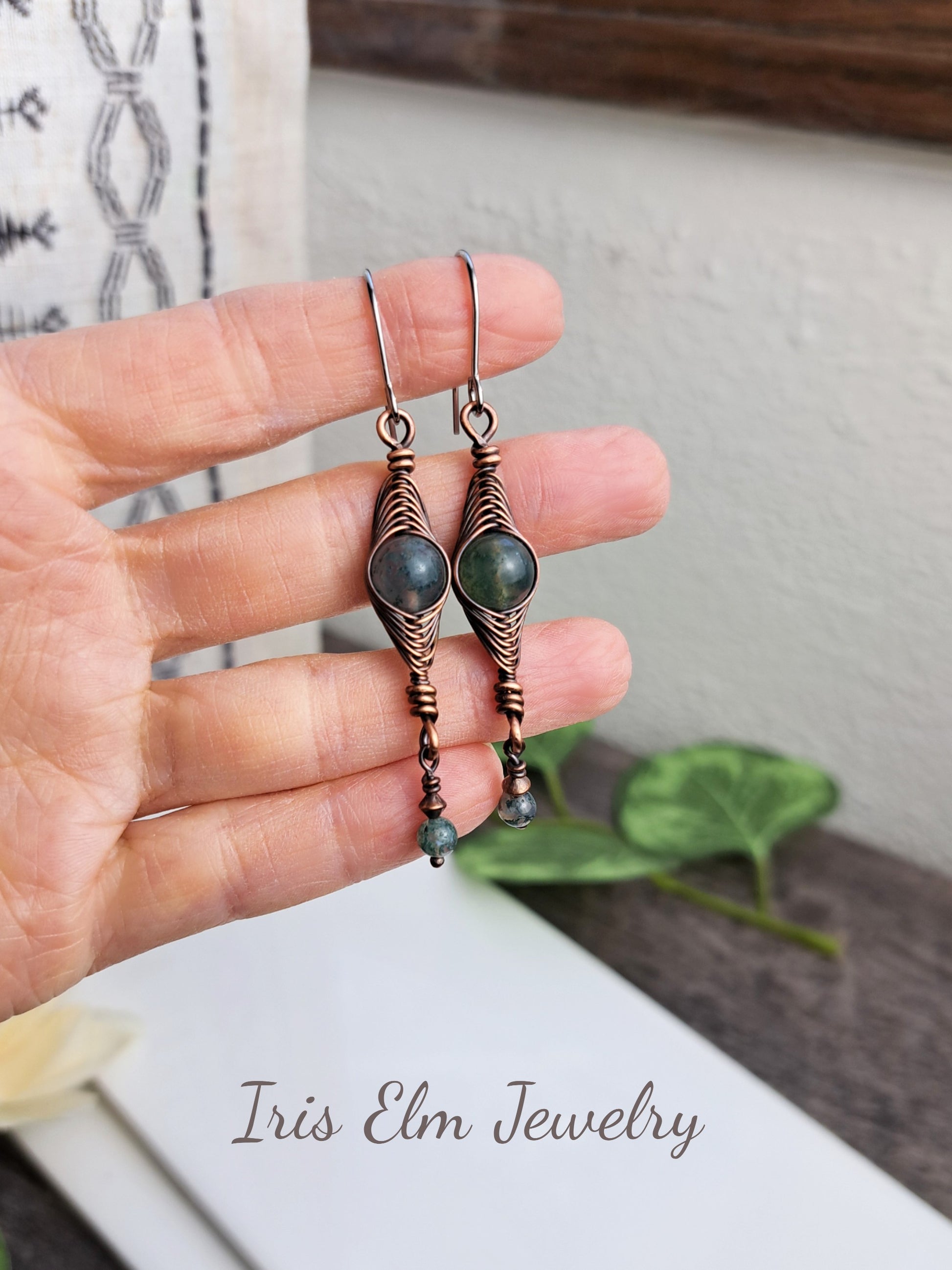 Hand holding a pair of copper wire wrapped moss agate herringbone earrings with 'Iris Elm Jewelry' branding.