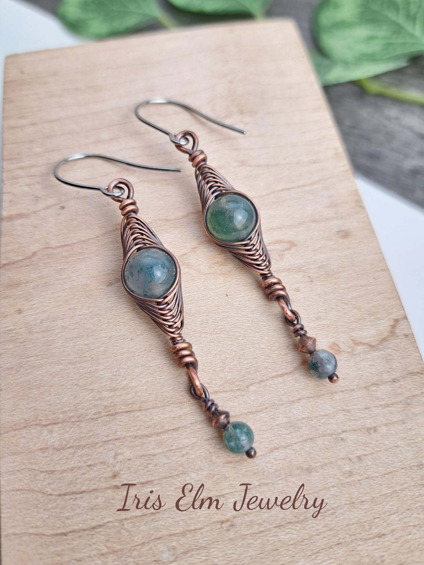 Pair of copper wire-wrapped herringbone earrings with green moss agate gemstones on a wooden surface, featuring 'Iris Elm Jewelry'.