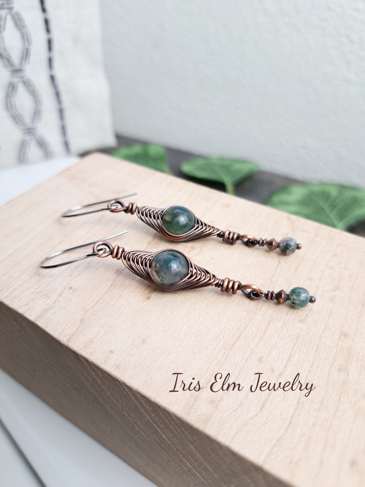Pair of handmade copper wire wrapped earrings with green moss agate gemstones on a wooden surface, branded 'Iris Elm Jewelry'. 