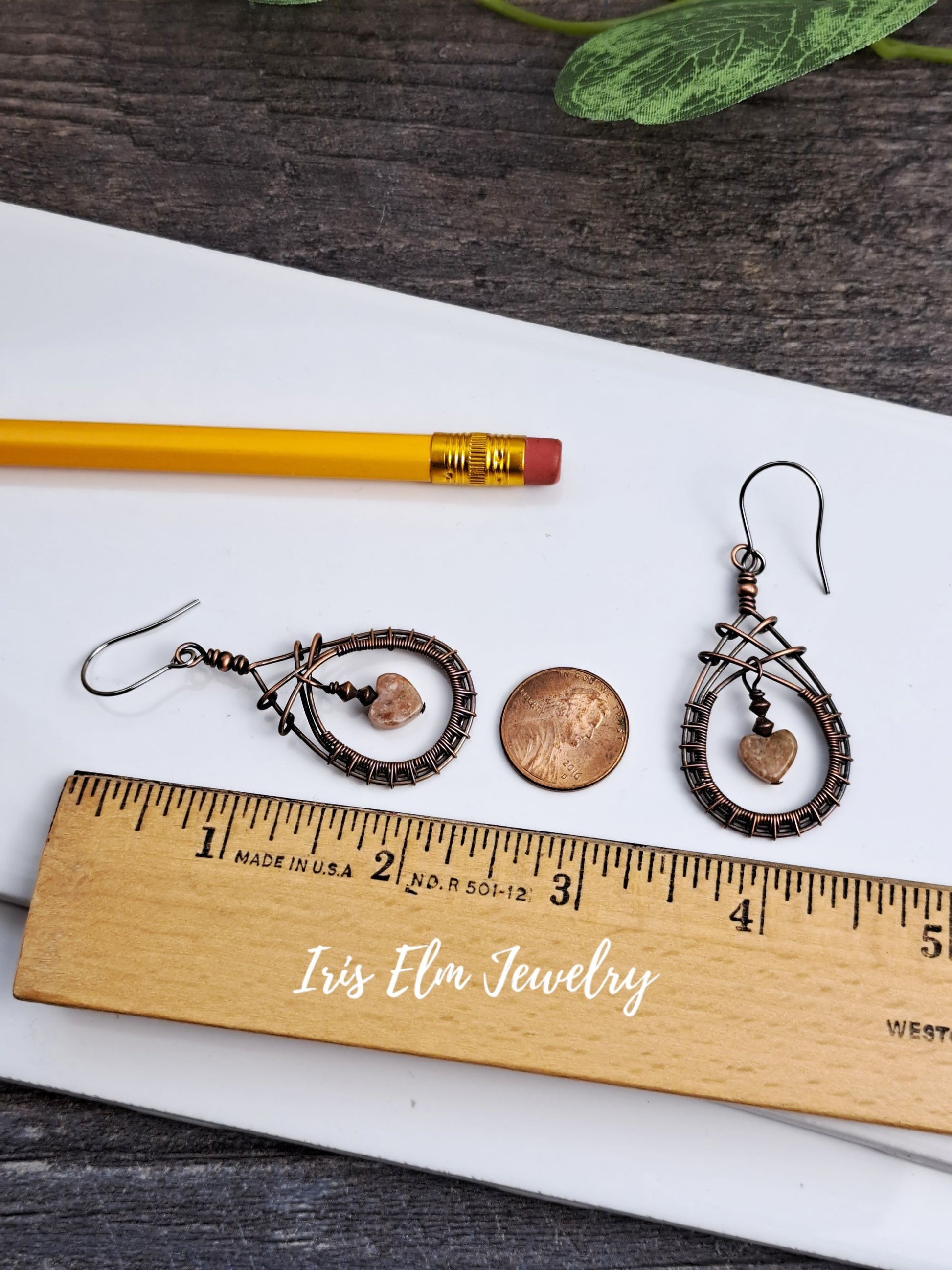 Pair of 2.25 inch long copper wire wrapped teardrop jasper earrings with heart-shaped stones on a wooden ruler, pencil, and white paper with green leaves in the background.