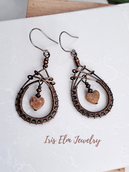 Pair of handmade copper wire-wrapped earrings with heart-shaped stones on a white surface, branded 'Iris Elm Jewelry'.