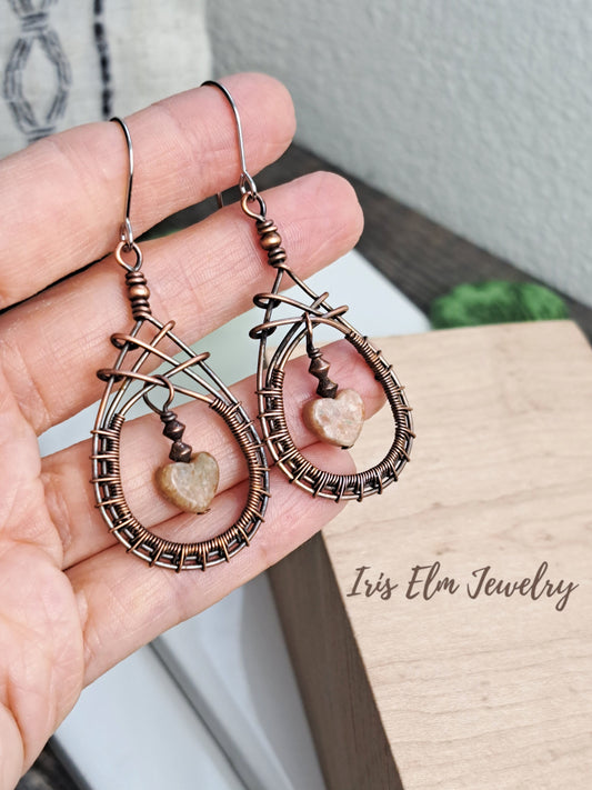 Hand holding copper wire-wrapped earrings with autumn jasper heart-shaped stones, branded 'Iris Elm Jewelry'.