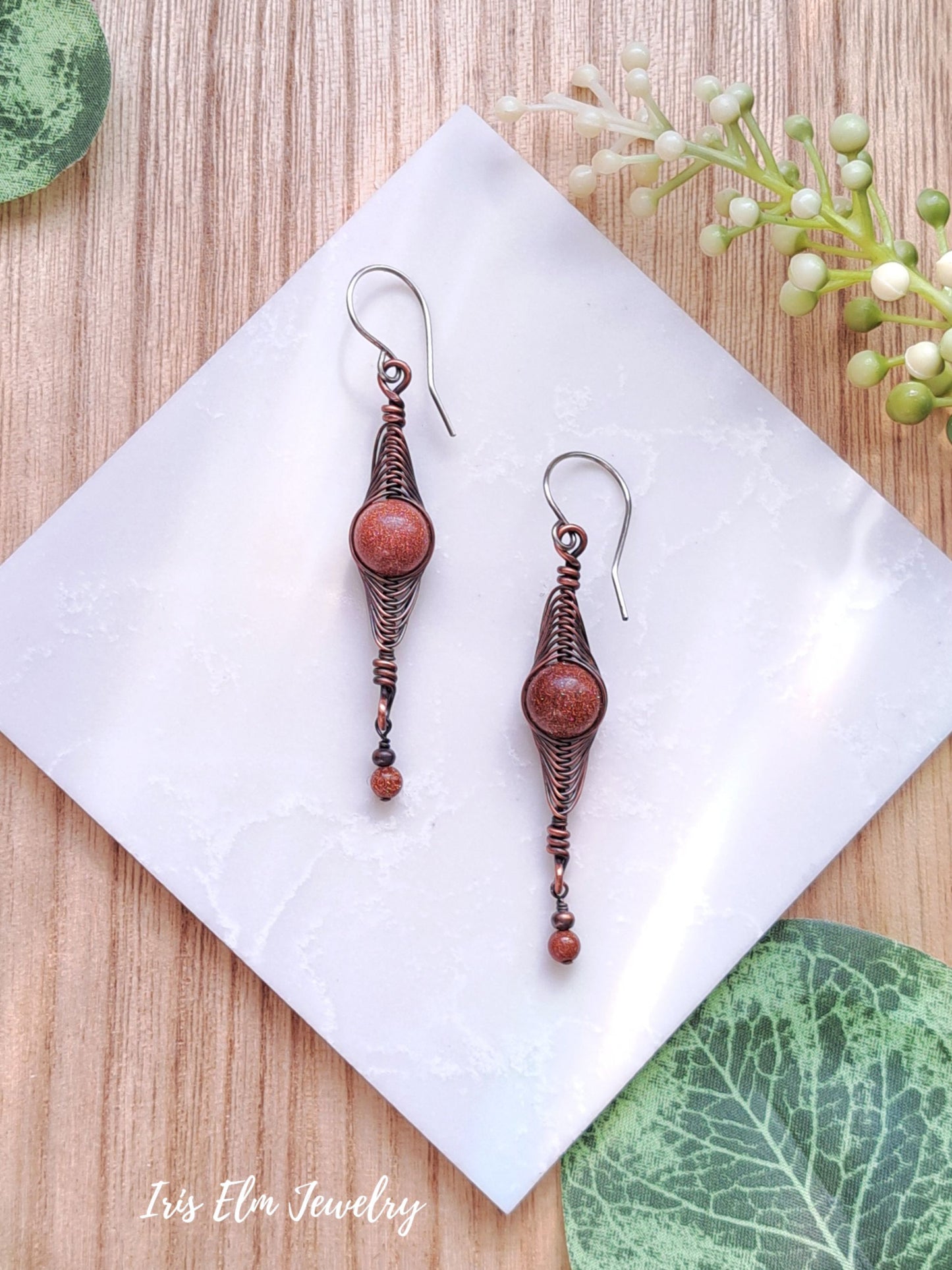 Pair of copper earrings with brown goldstone beads on a white napkin with green leaves in the background.