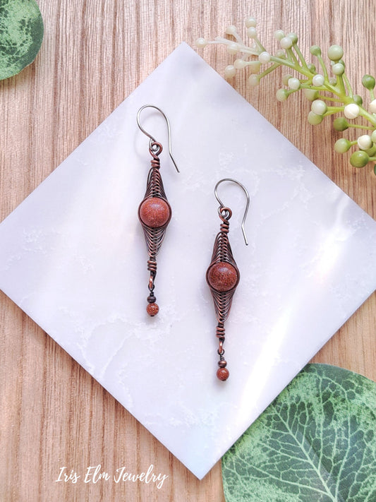 Pair of copper earrings with brown goldstone beads on a white napkin with green leaves in the background.