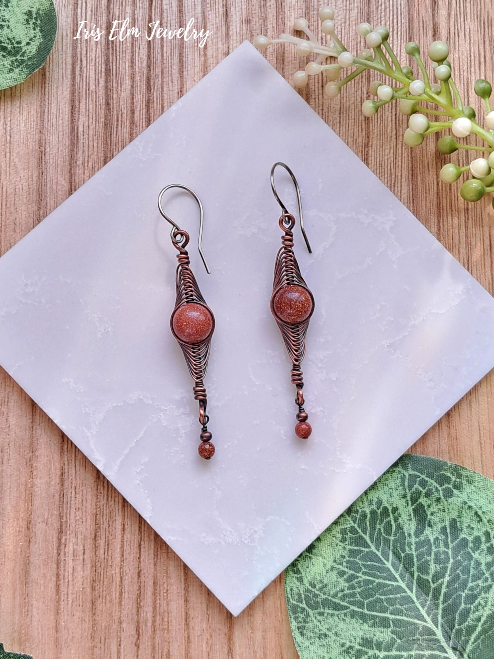 Pair of copper wire wrapped herringbone earrings with brown goldstones on a white napkin with green leaves in the background.