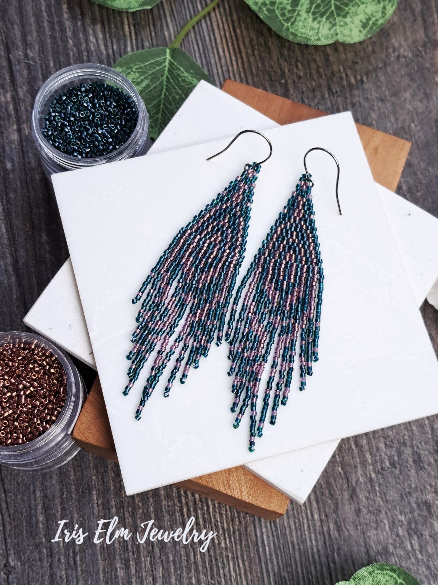 Beaded forest green and pink seed bead fringe art deco earrings on a white card with 'Iris Elm Jewelry' branding, surrounded by small containers of beads.