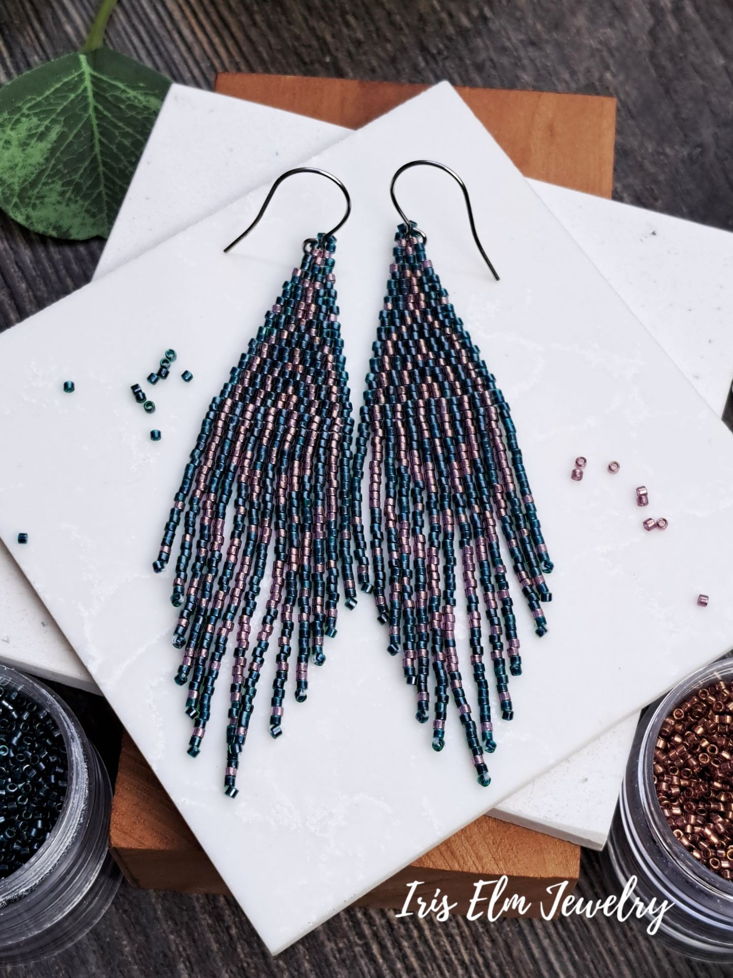 Beaded forest green and pink seed bead fringe art deco earrings on a white marble surface with beads and a wooden block.