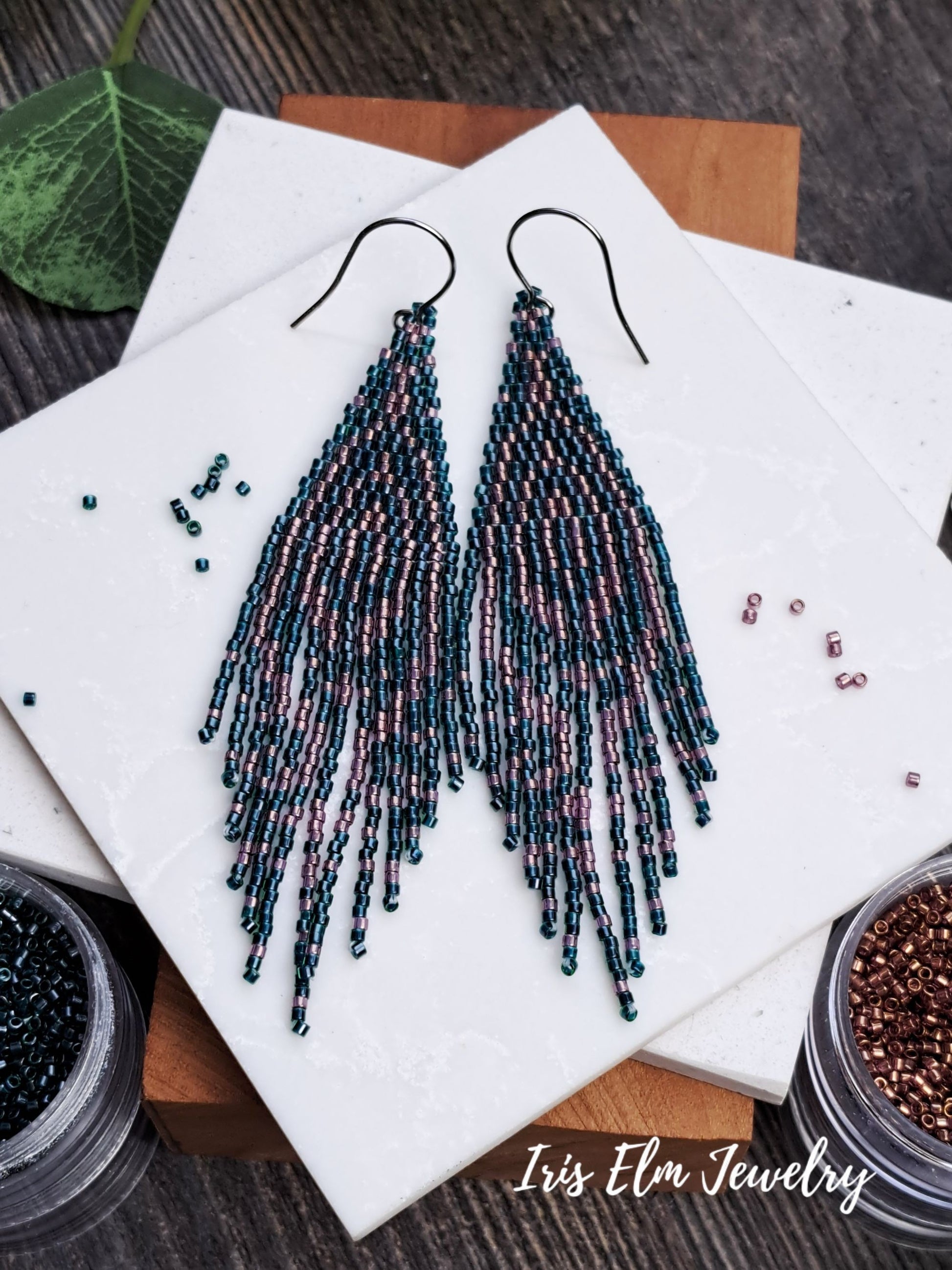 Beaded forest green and pink seed bead fringe art deco earrings on a white marble surface with beads and a wooden block.
