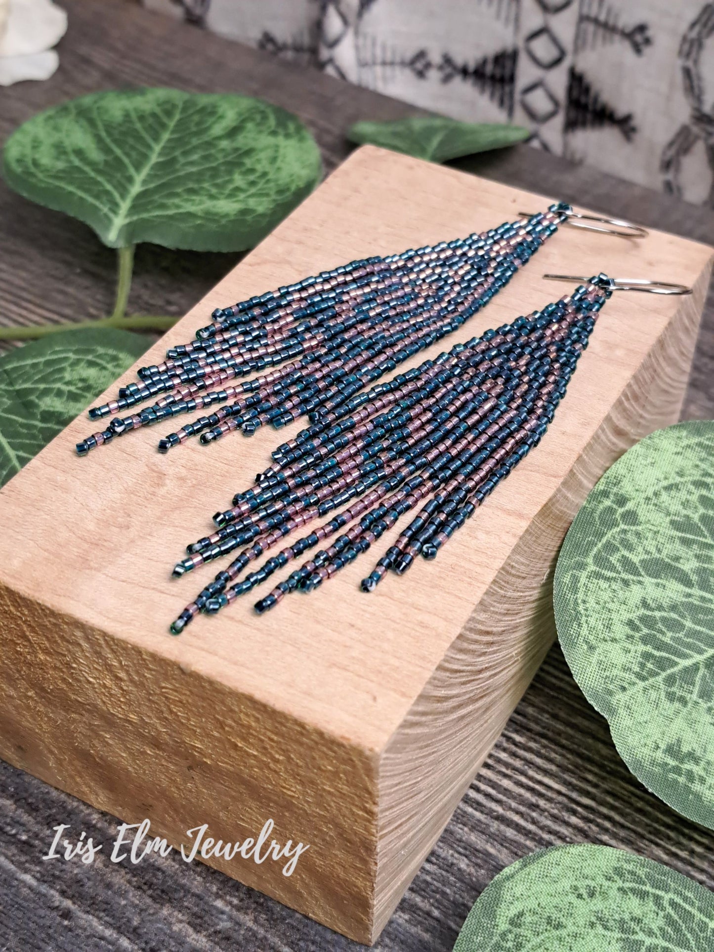 Beaded forest green and pink seed bead fringe art deco earrings on a wooden block with green leaves in the background, featuring 'Iris Elm Jewelry'.