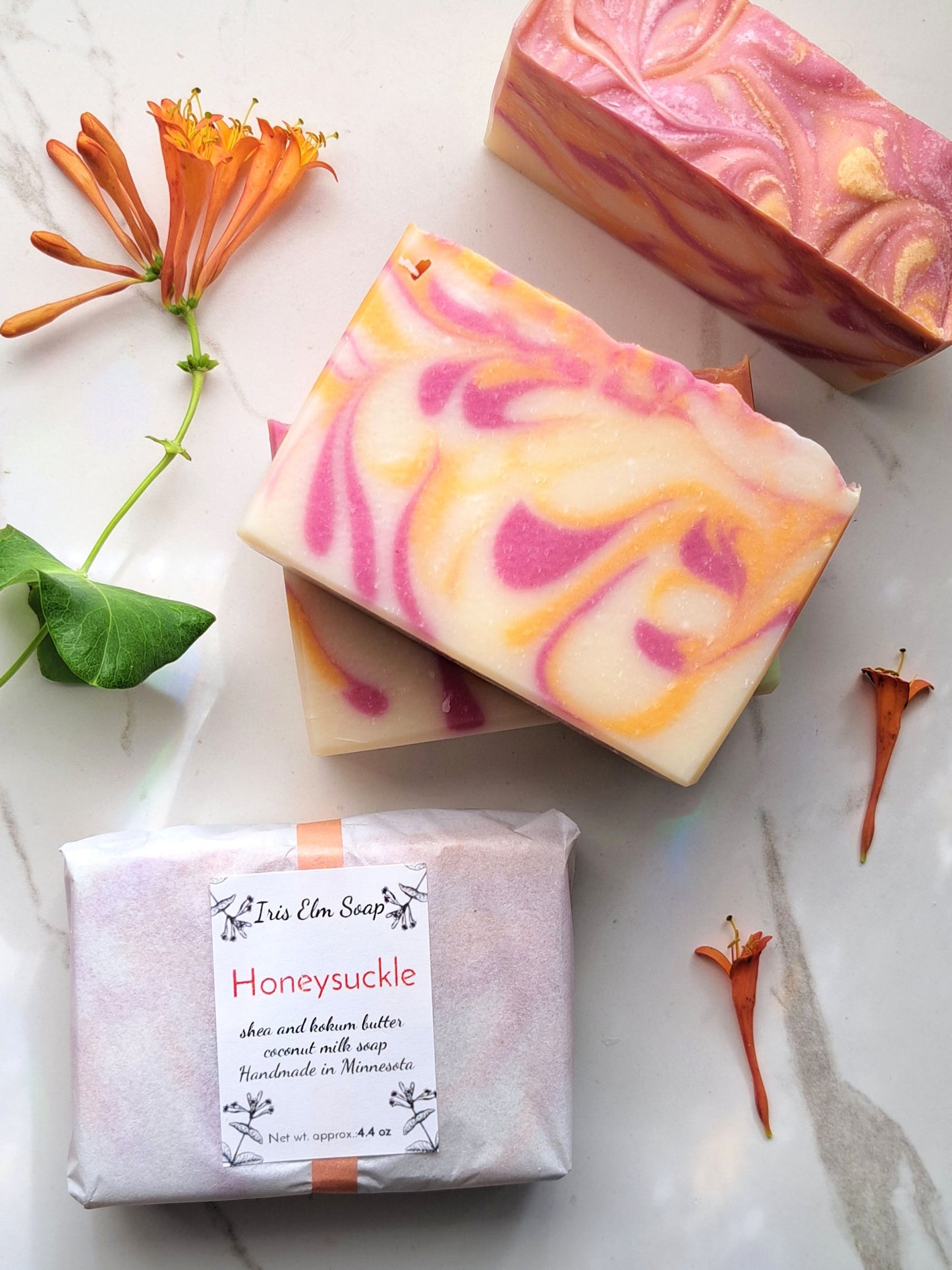 Three bars of honeysuckle handmade soap with pink and orange swirl pattern on a white surface with flowers and leaves.