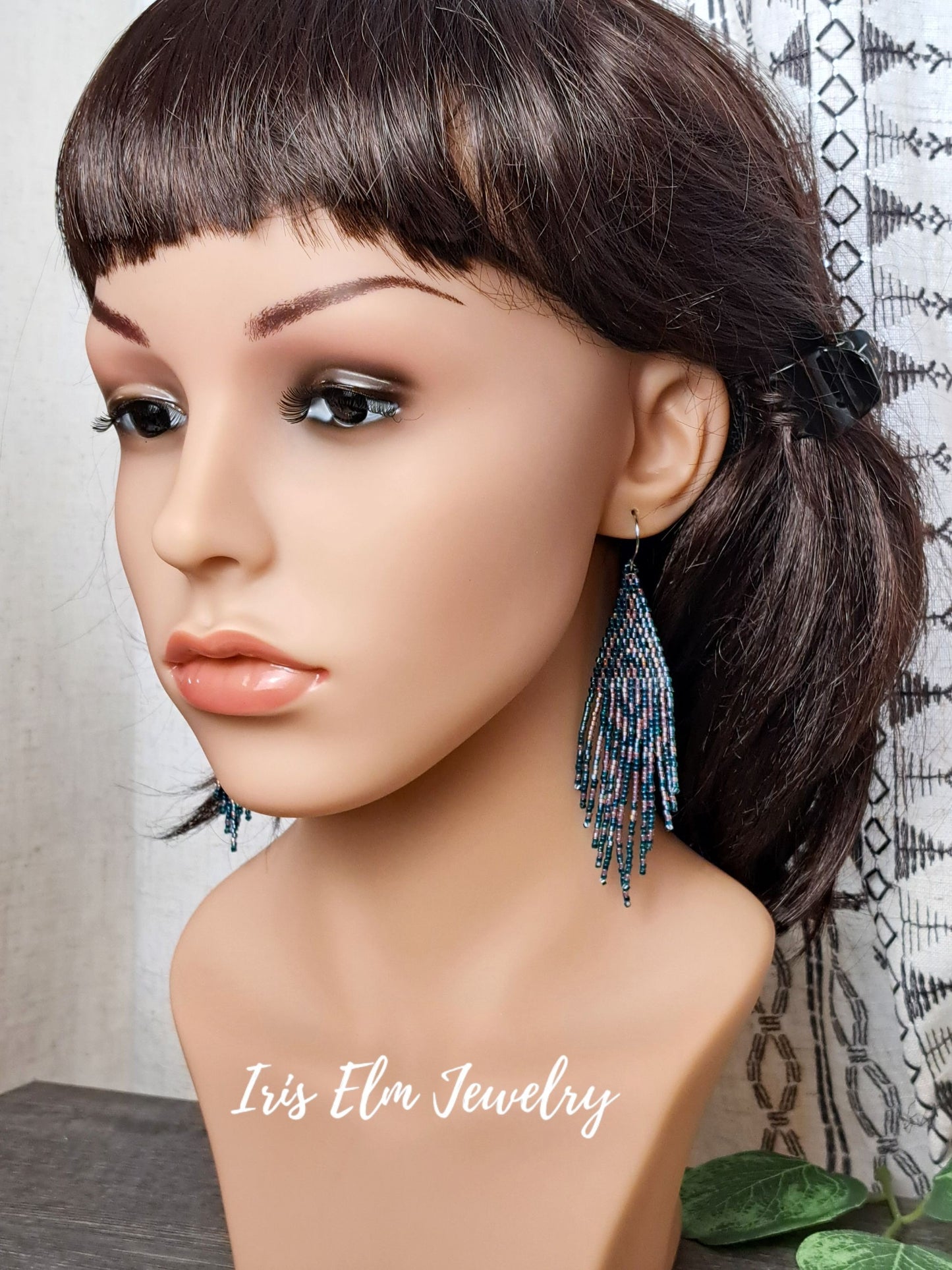 Mannequin head with dark hair styled in a ponytail wearing forest green and pink seed bead fringe art deco beaded earrings, with 'Iris Elm Jewelry' branding.