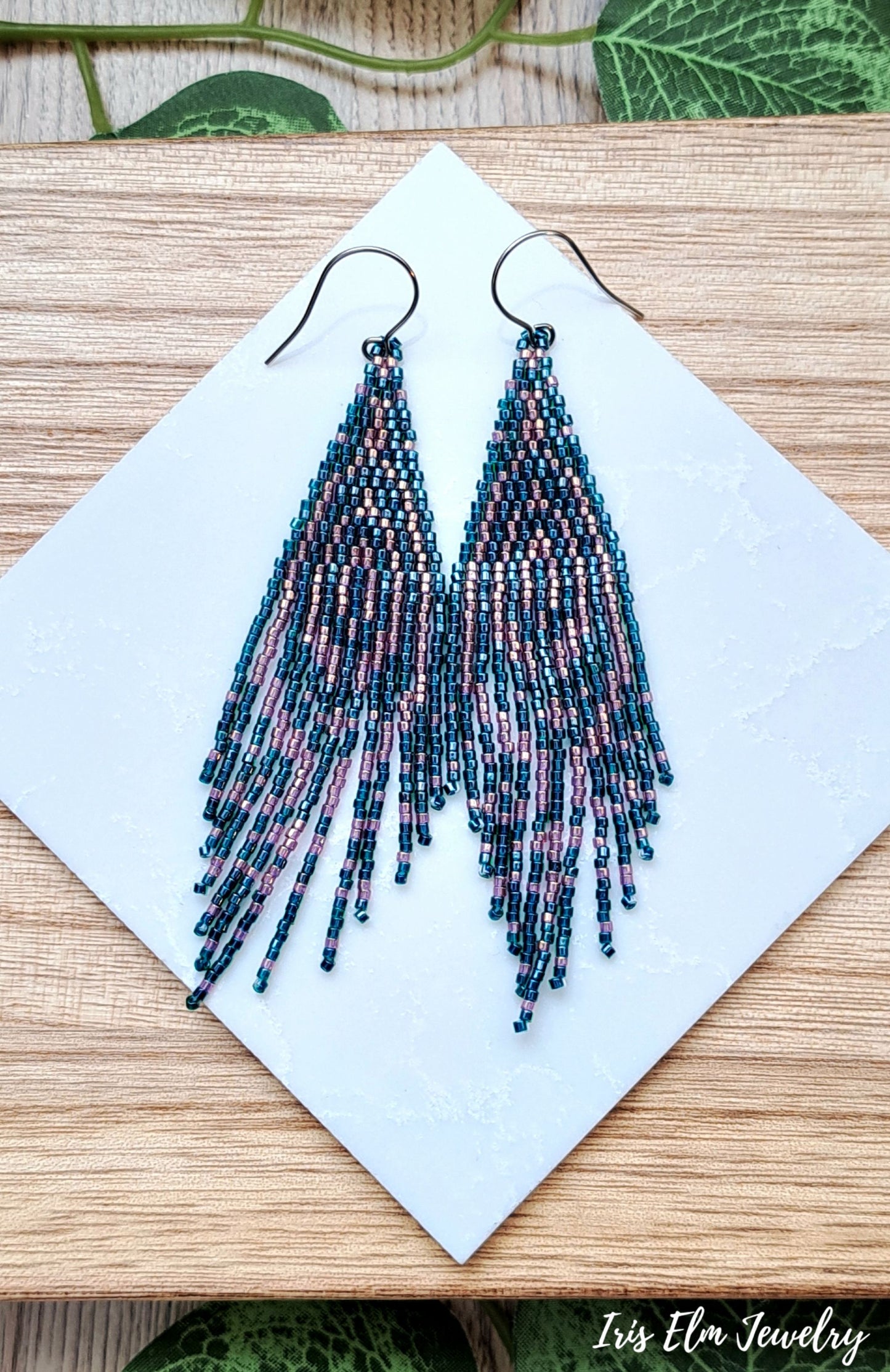 Beaded forest green and pink seed bead fringe art deco earrings on a white card with a wooden background