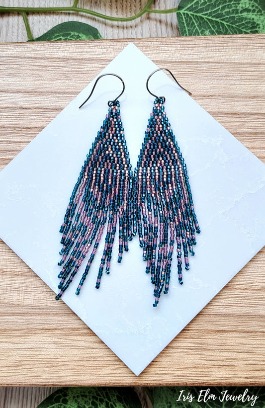 Beaded forest green and pink seed bead fringe art deco earrings on a white card with a wooden background