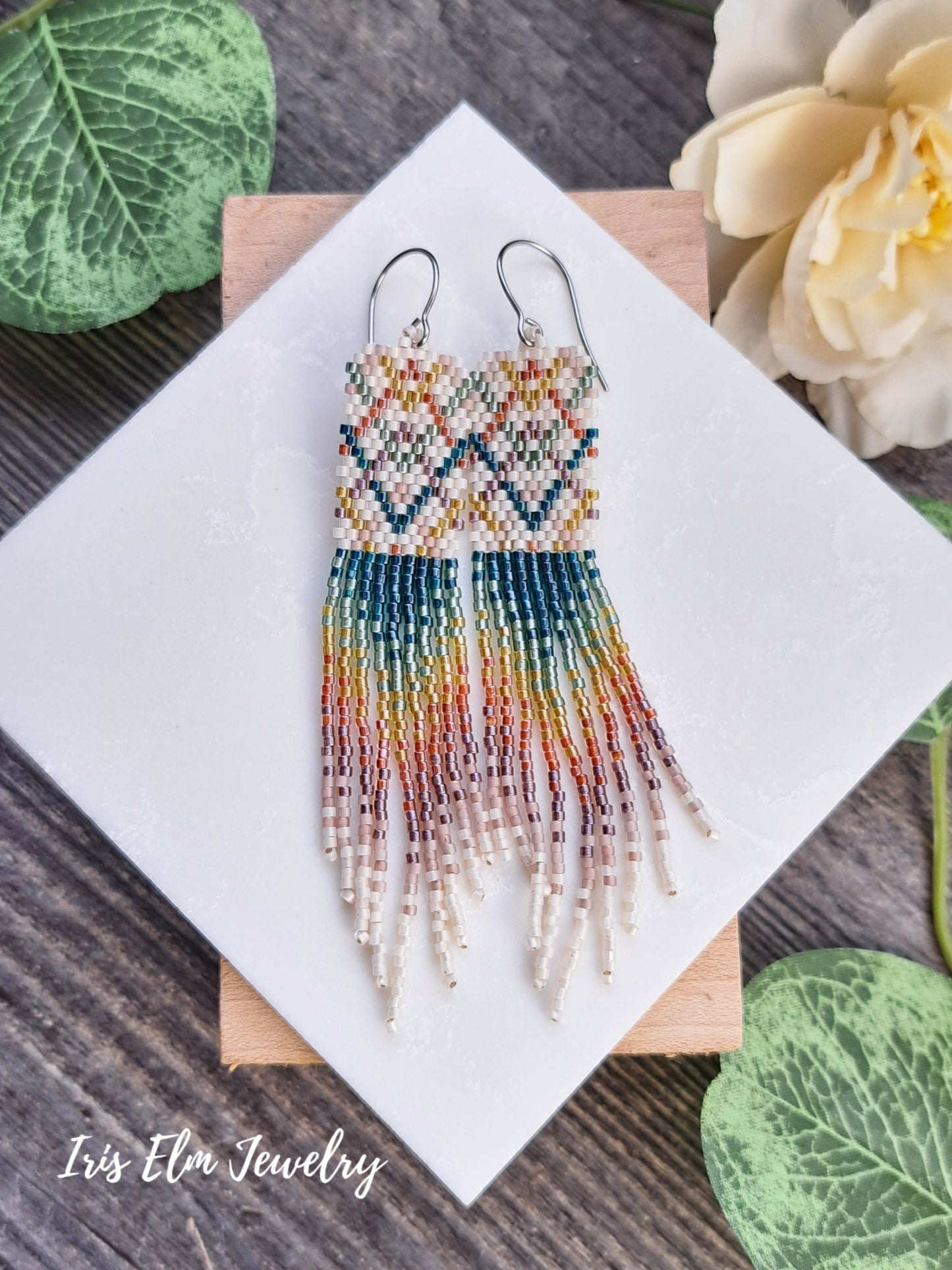 Colorful beaded plaid rainbow seed bead fringe earrings on a white square with 'Iris Elm Jewelry' branding, surrounded by green leaves and flowers.