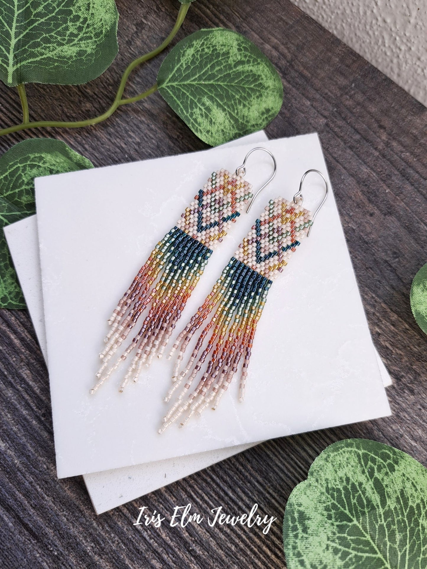 Colorful beaded plaid rainbow seed bead fringe handmade beadwork earrings on a white card with green leaves in the background