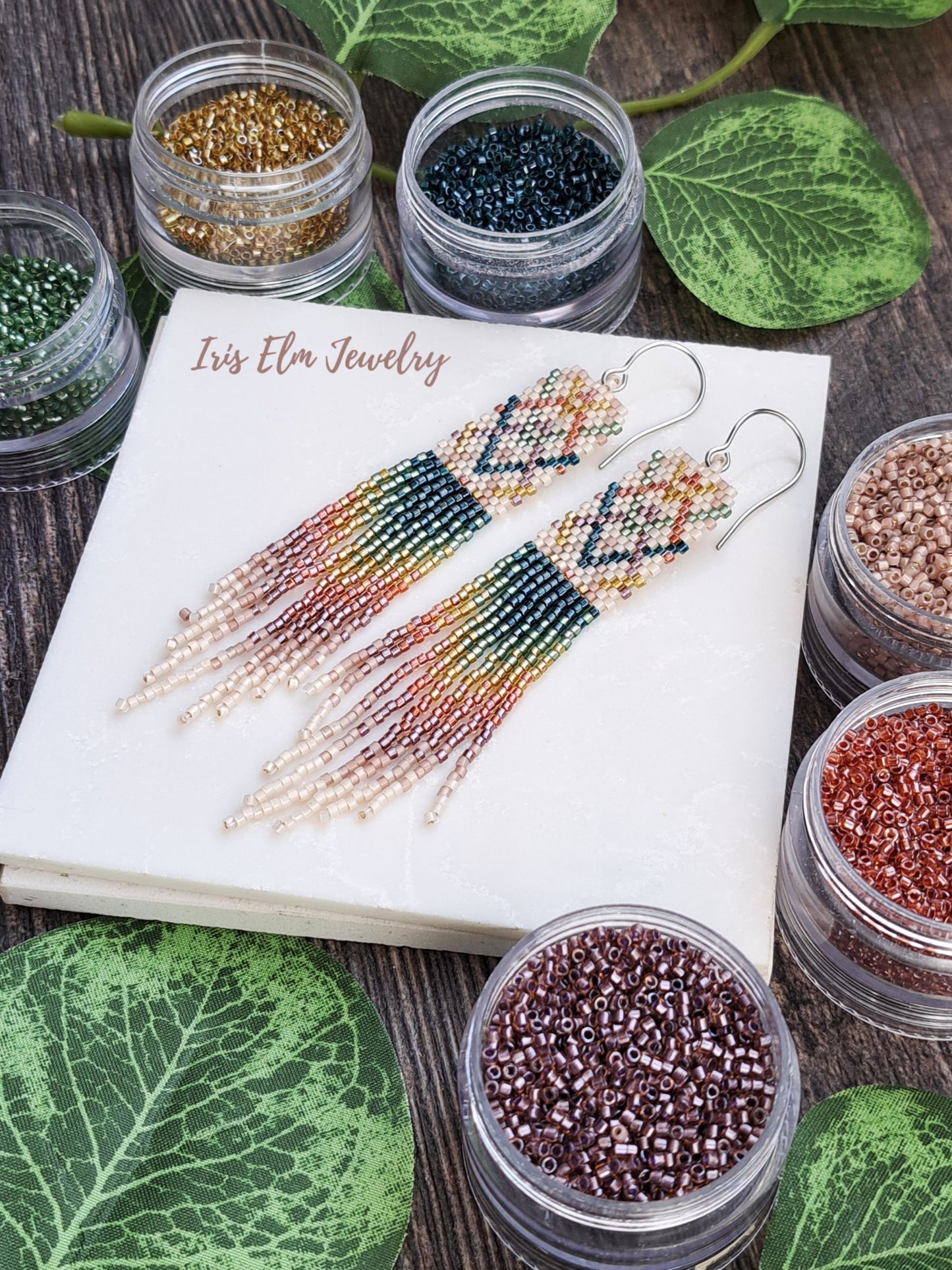 Beaded plaid rainbow seed bead fringe earrings on a notebook with containers of beads and green leaves around