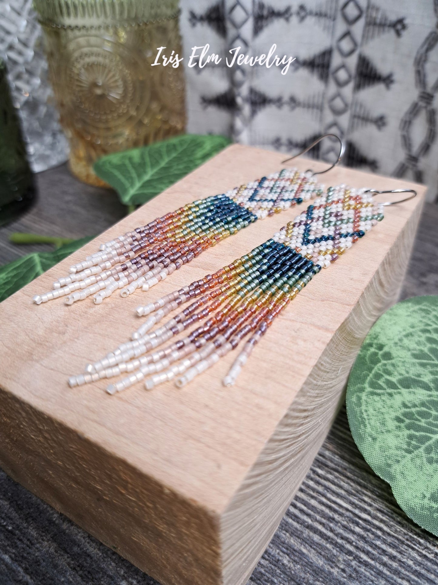Colorful beaded plaid rainbow seed bead fringe earrings on a wooden block with 'Iris Elm Jewelry' branding.