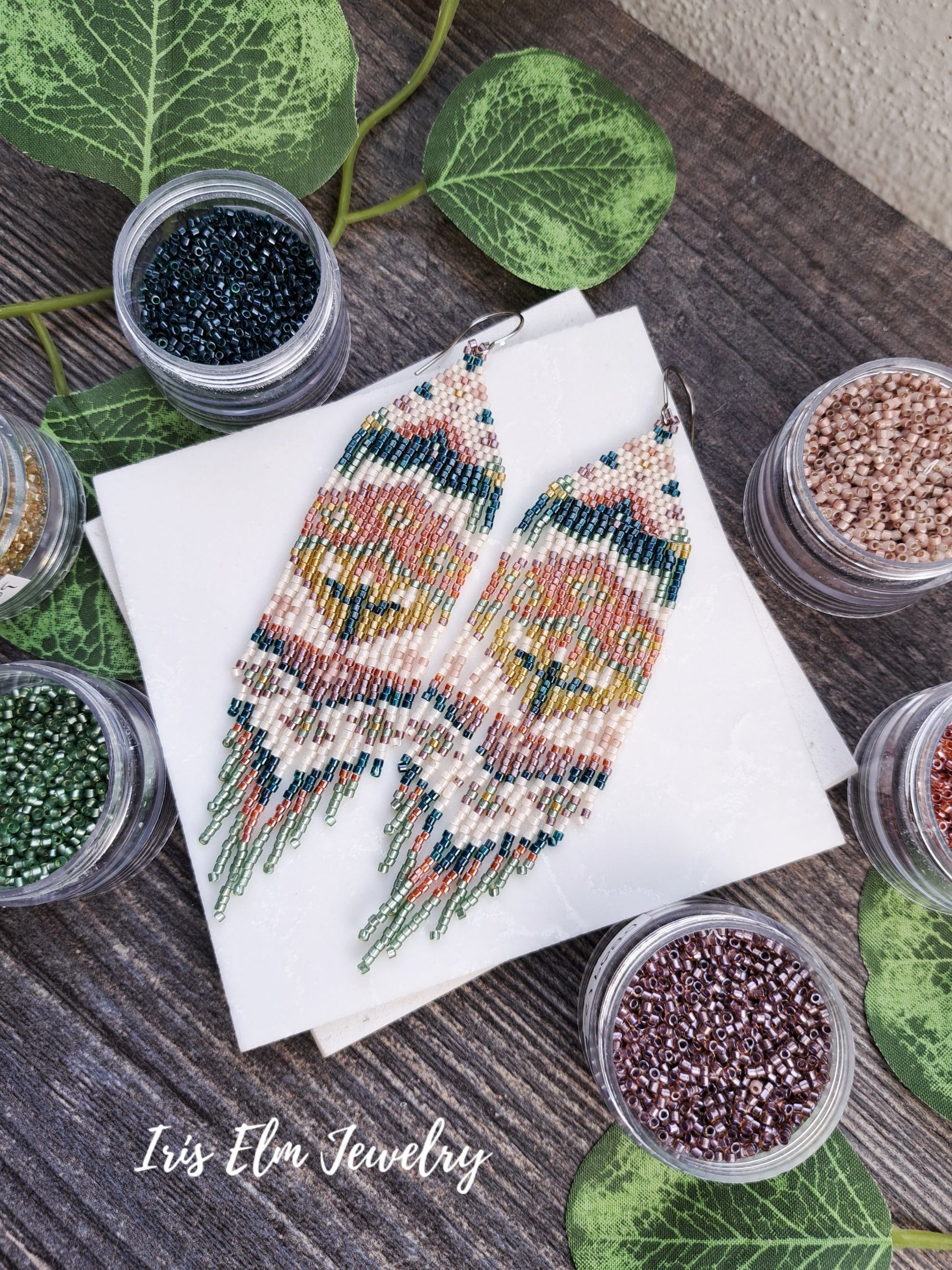 handmade beadwork green, orange, yellow, pink spring floral folk art seed bead fringe earrings on a book with containers of beads around, on a wooden surface.