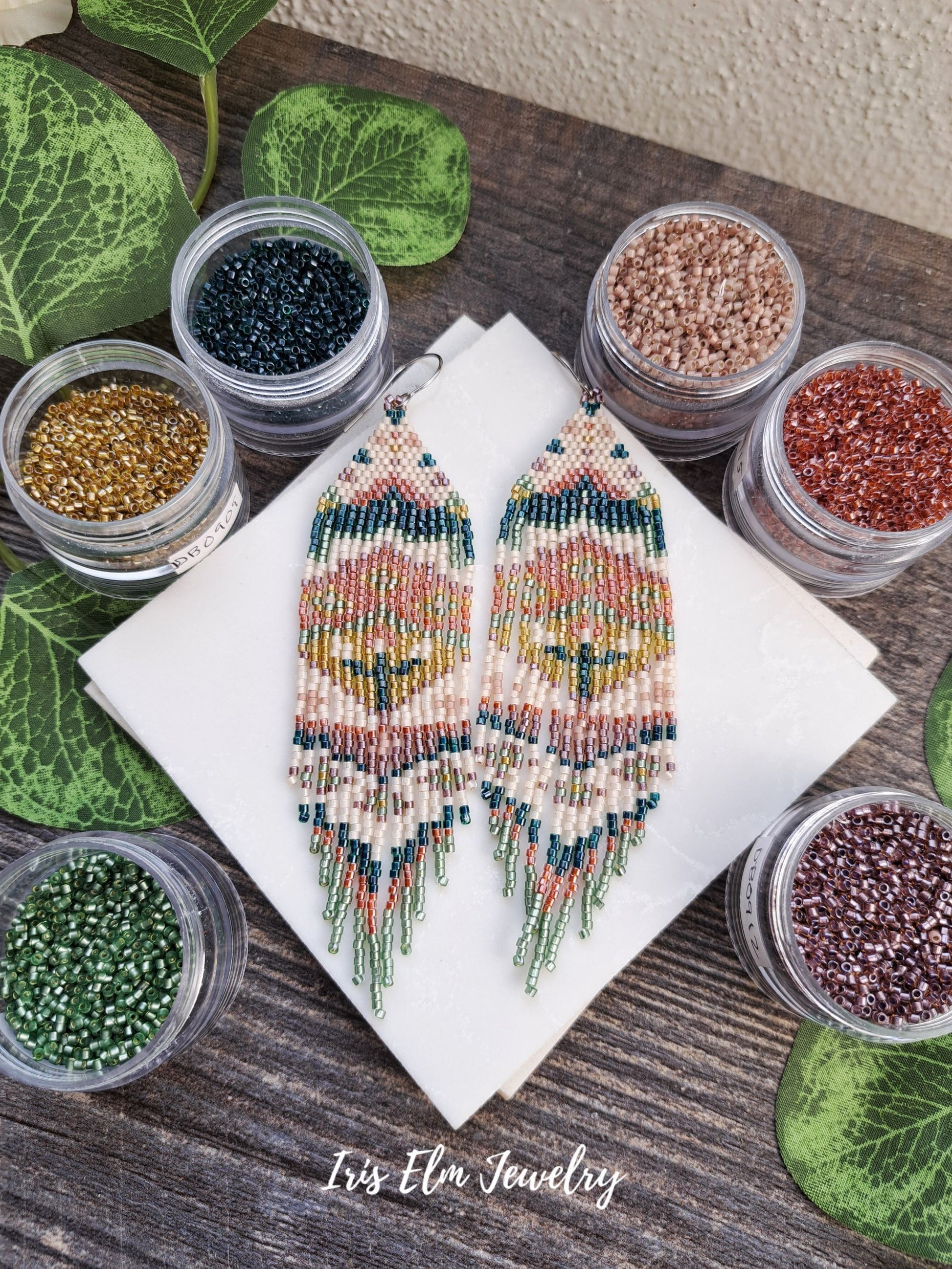 Beaded green, orange, yellow, pink spring floral folk art seed bead fringe earrings on a wooden surface with small containers of beads and 'Iris Elm Jewelry' branding.