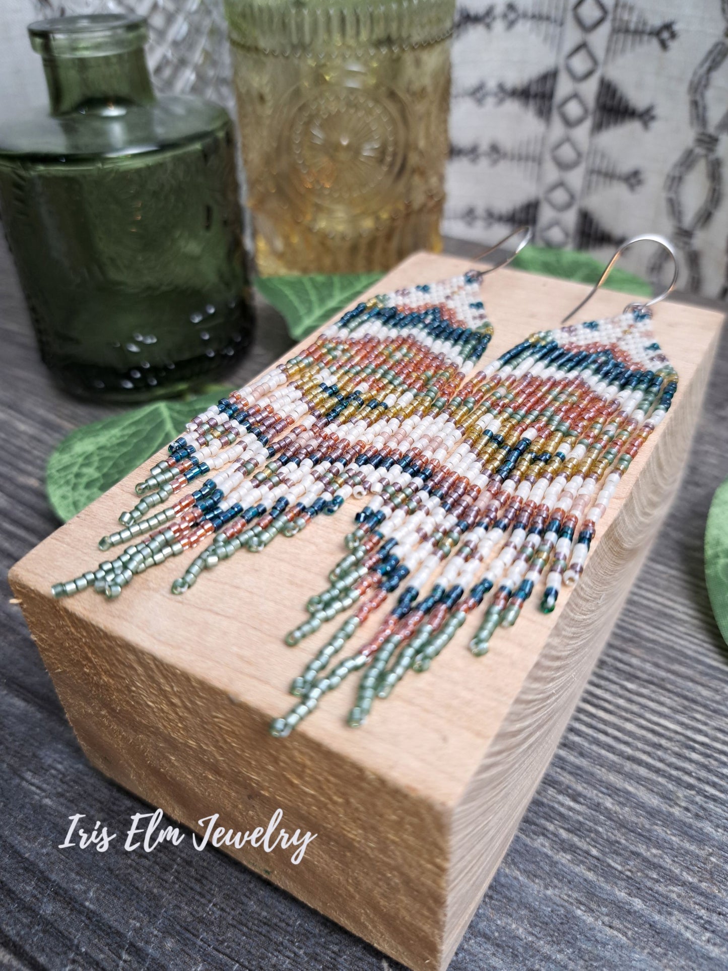 green, orange, yellow, pink spring floral folk art seed bead fringe earrings on a wooden block with 'Iris Elm Jewelry' branding.