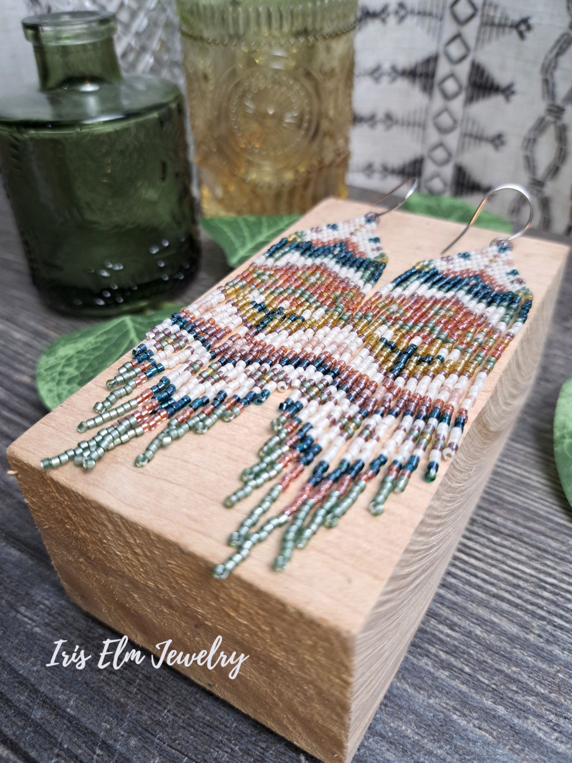 green, orange, yellow, pink spring floral folk art seed bead fringe earrings on a wooden block with 'Iris Elm Jewelry' branding.