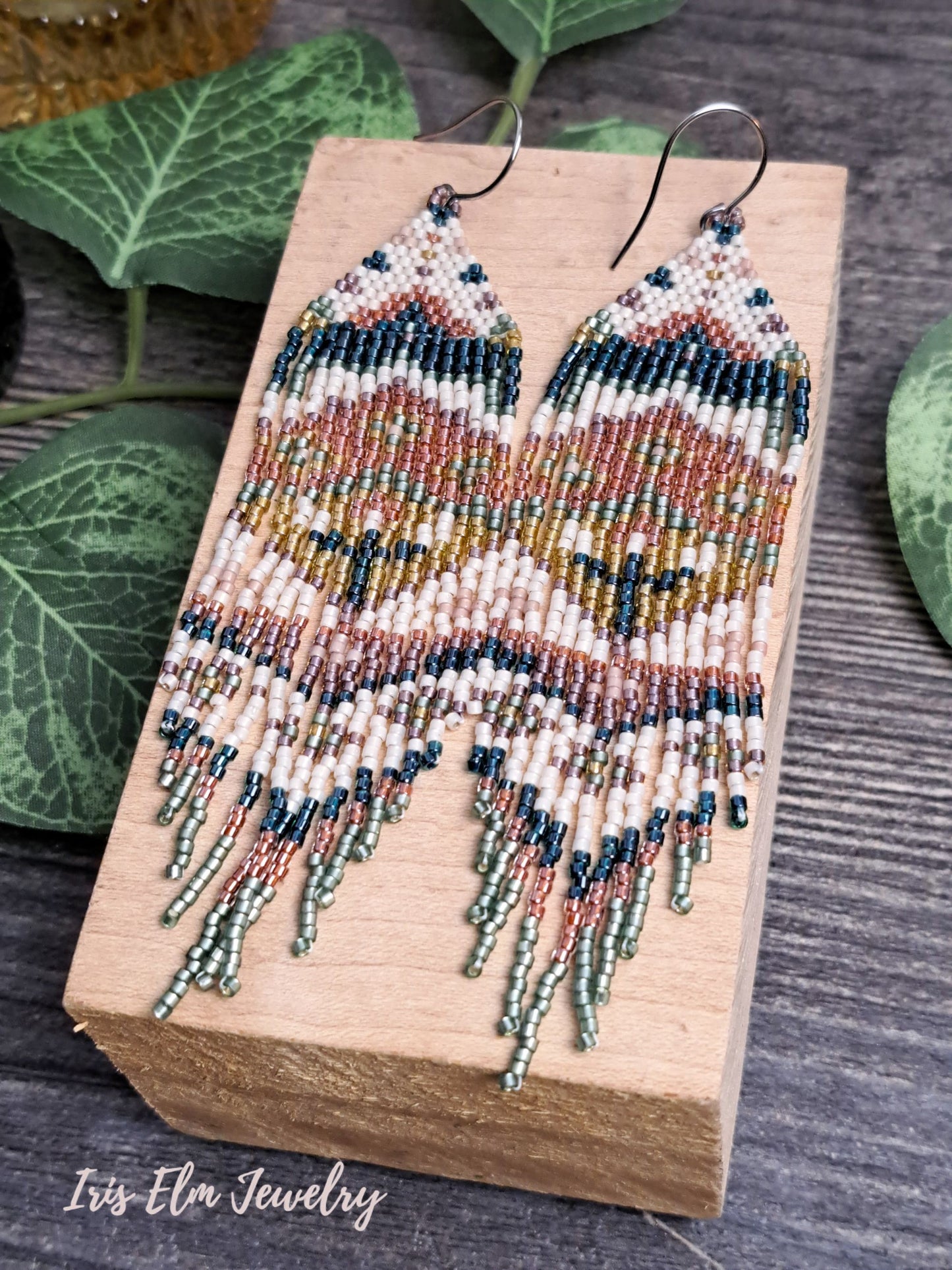 Beaded green, orange, yellow, pink spring floral folk art seed bead fringe earrings on a wooden block with green leaves in the background