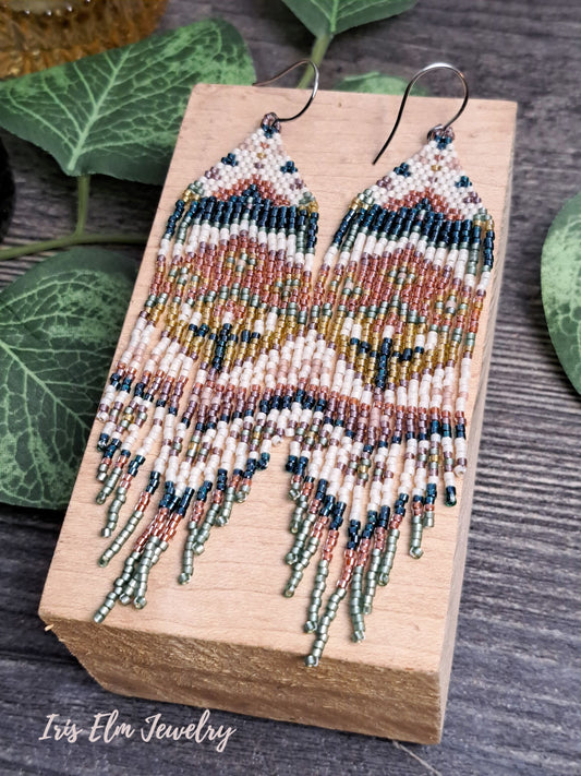 Beaded green, orange, yellow, pink spring floral folk art seed bead fringe earrings on a wooden block with green leaves in the background