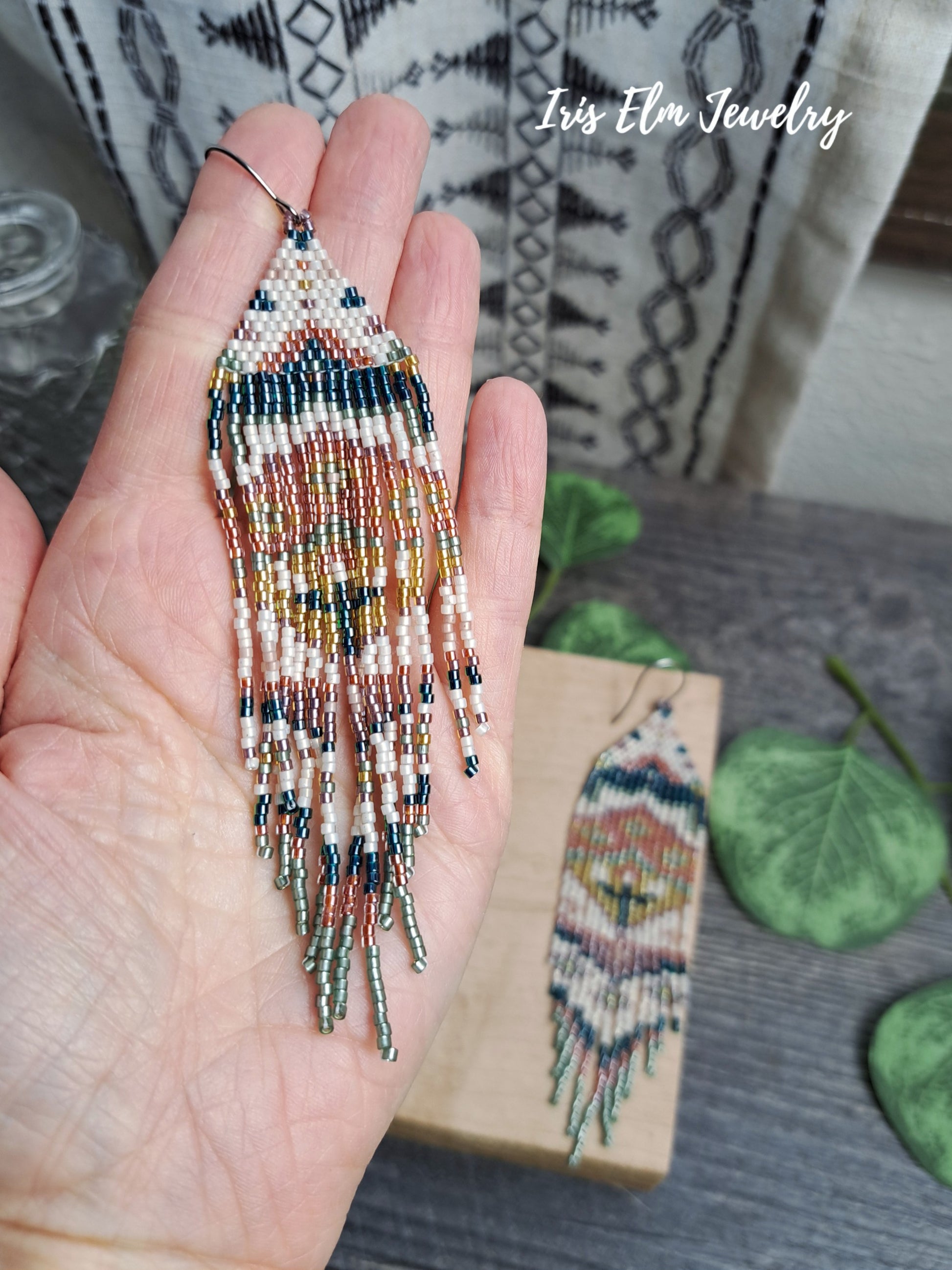 Beaded green, orange, yellow, pink spring floral folk art seed bead fringe earrings held in a hand with a blurred background