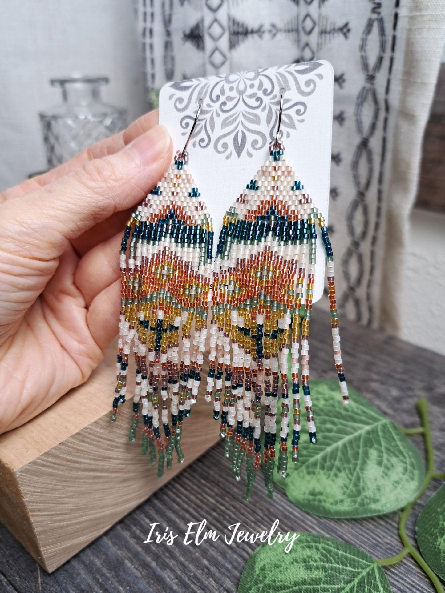 Hand holding beaded green, orange, yellow, pink spring floral folk art seed bead fringe earrings on a card with 'Iris Elm Jewelry' branding.