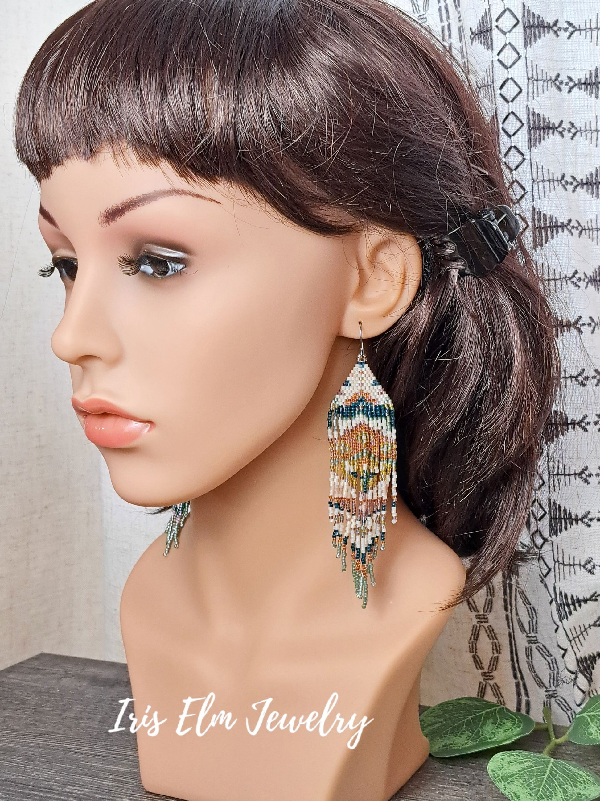 Mannequin head with beaded green, orange, yellow, pink spring floral folk art seed bead fringe earrings on a neutral background, featuring 'Iris Elm Jewelry'.