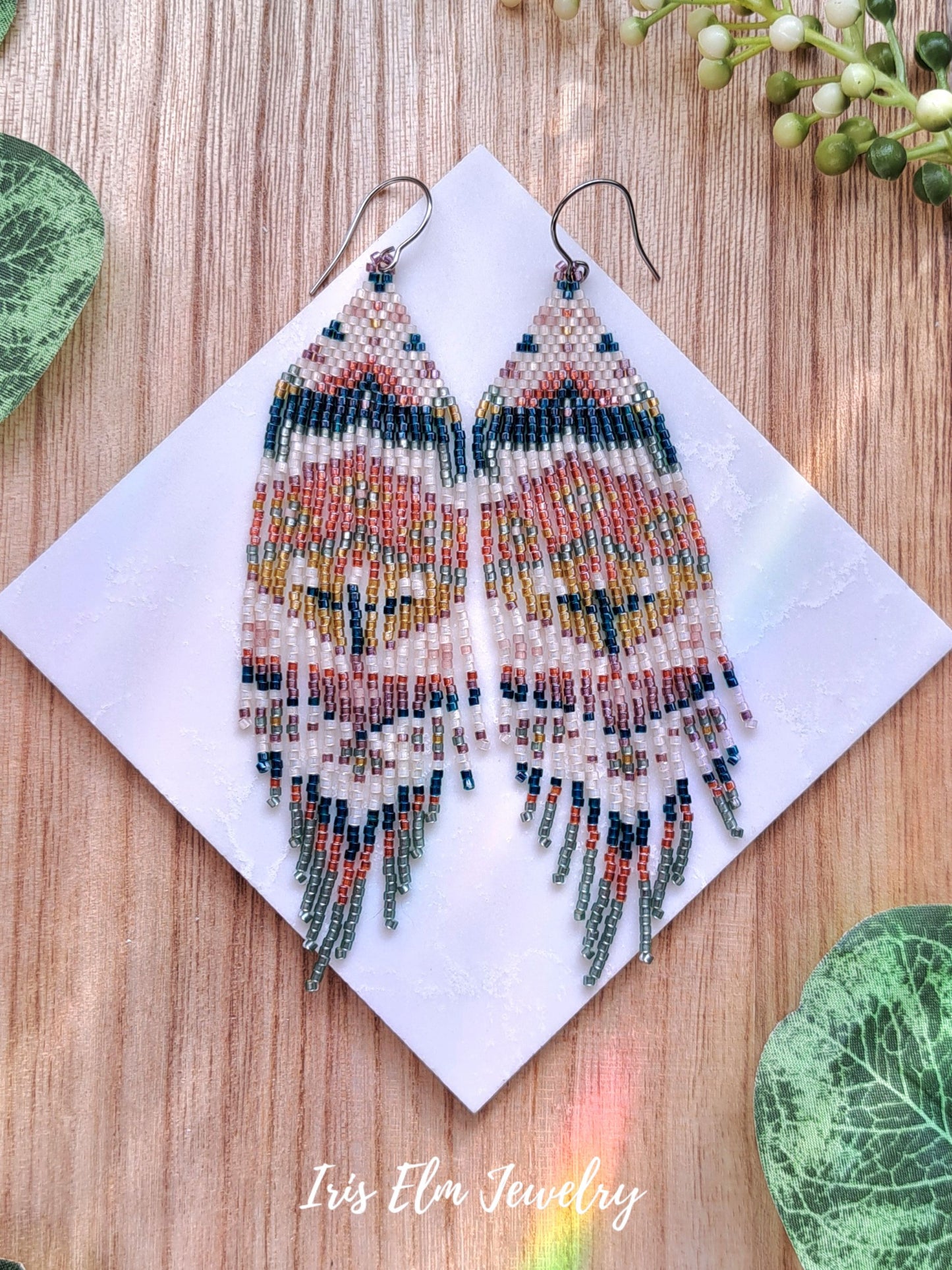 Colorful beaded spring floral folk art seed bead fringe earrings on a wooden surface with 'Iris Elm Jewelry' branding.