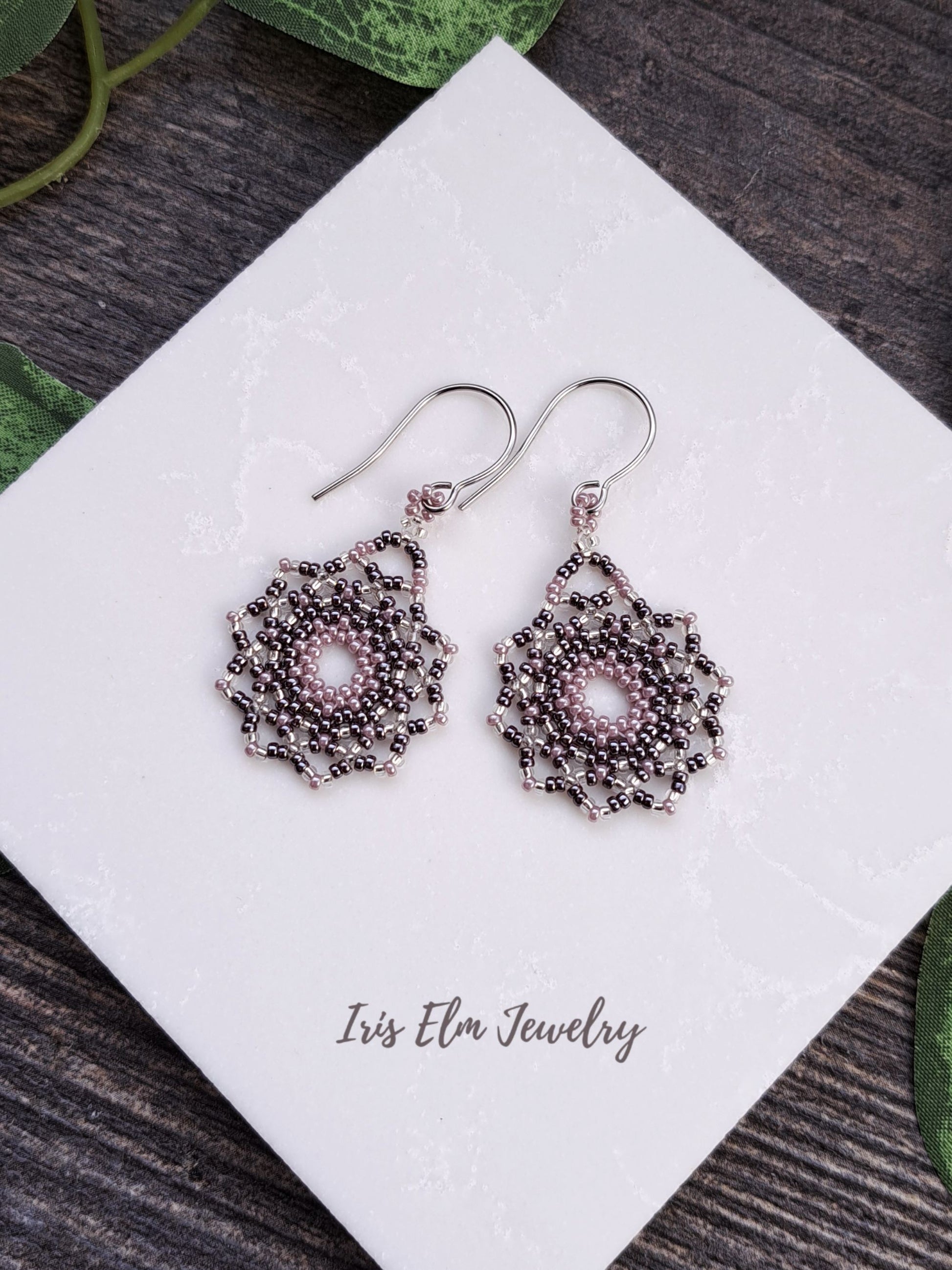 Decorative lilac purple seed bead mandala earrings on a white card with 'Iris Elm Jewelry' branding, placed on a wooden surface.