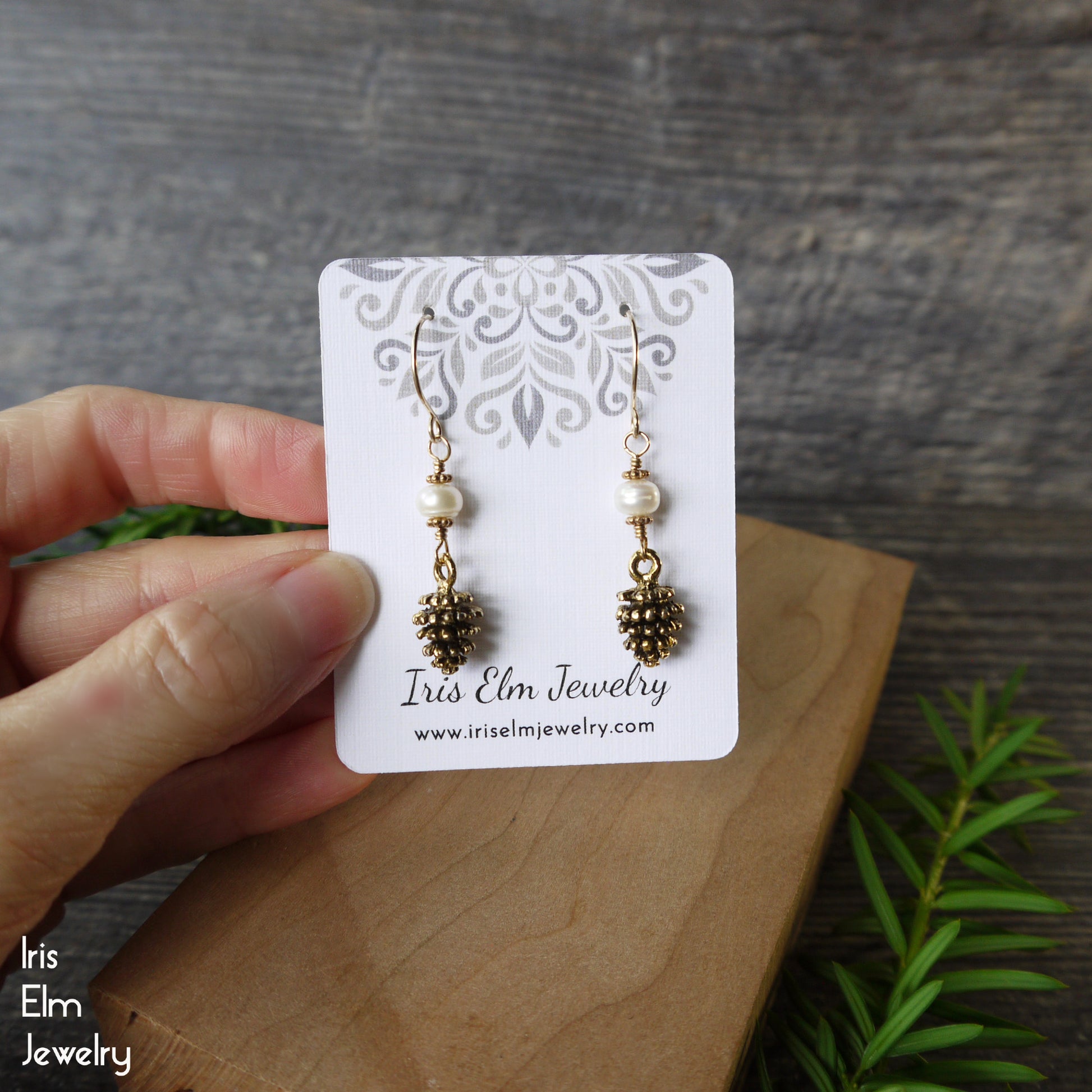 Pair of pinecone and freshwater pearl earrings on a 'Iris Elm Jewelry' card held by a hand with a wooden surface and greenery in the background.