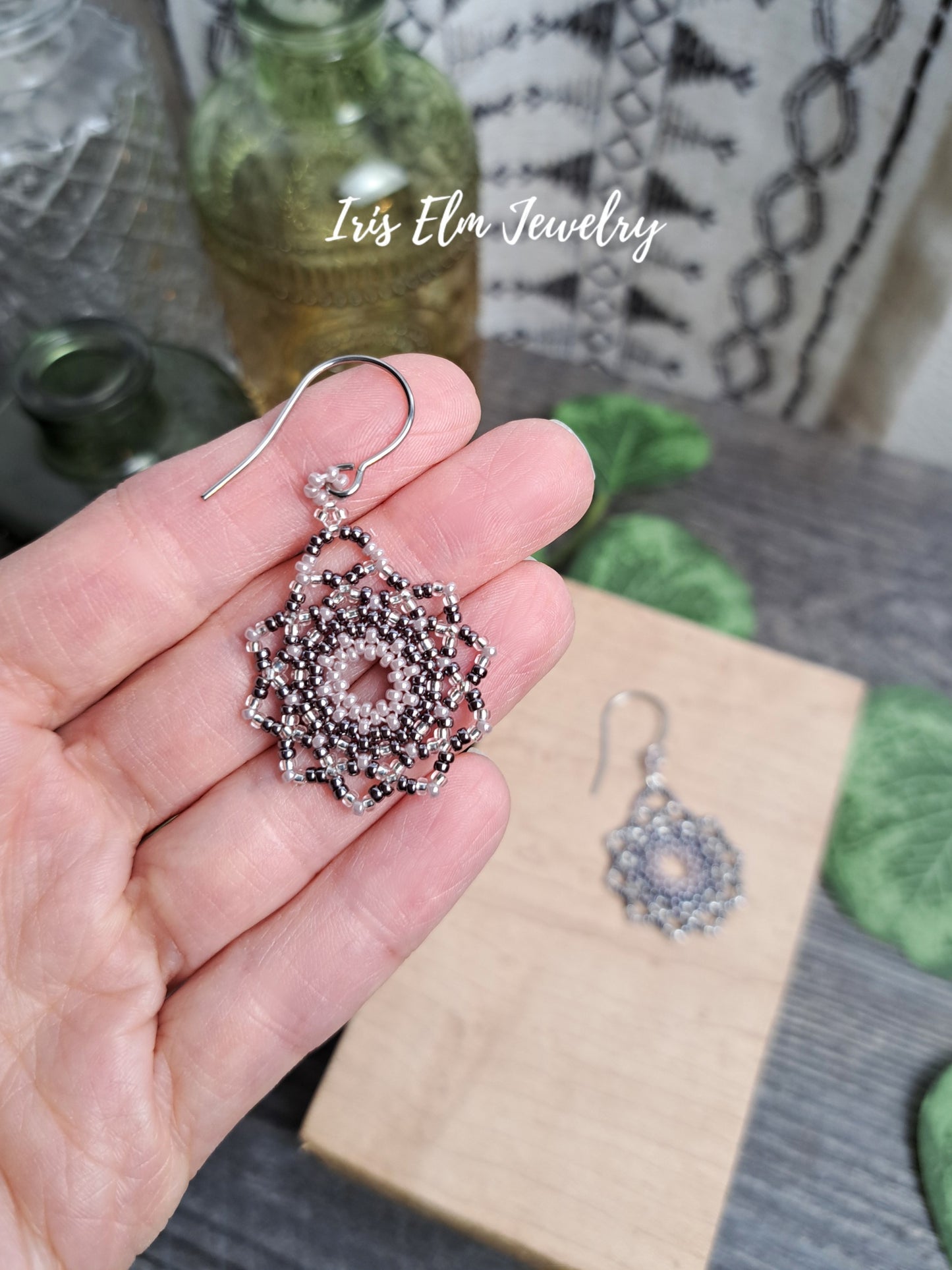 beadwoven lilac purple seed bead mandala earrings held in a hand with 'Iris Elm Jewelry' branding in the background.
