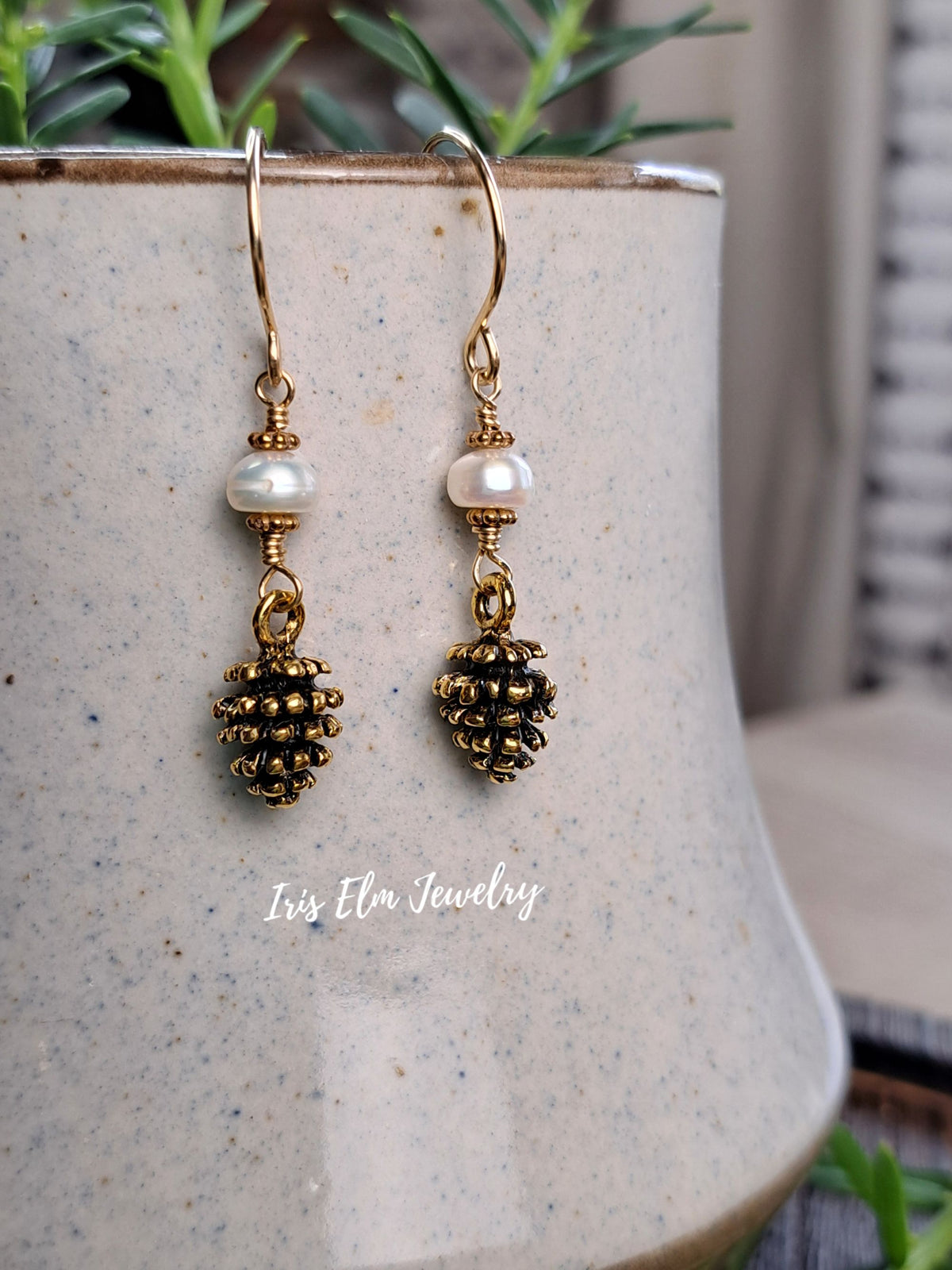 A pair of gold-toned dangle earrings with white pearl beads and textured pinecone charms, displayed on a speckled ceramic pot with green foliage in the background.