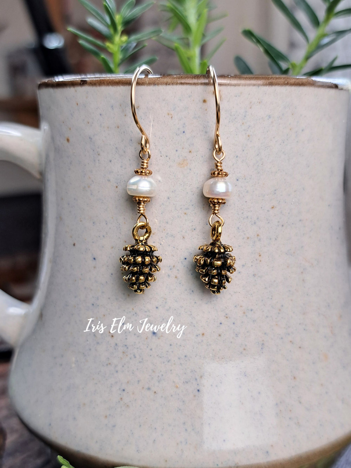 A pair of gold and pearl drop earrings with textured pinecone charms, displayed on a speckled ceramic mug with green plants in the background. The watermark 'Iris Elm Jewelry' is visible on the mug.