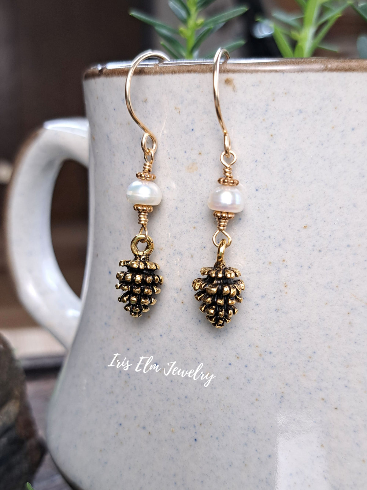 A pair of gold-toned dangle earrings with white pearl beads and pinecone charms, hanging in front of a speckled ceramic mug with a visible 'Iris Elm Jewelry' watermark.