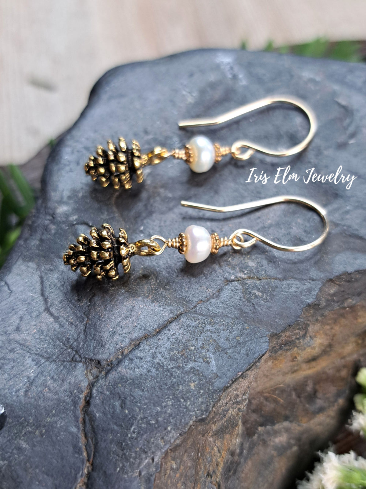 A pair of gold-tone dangle earrings with small pinecone charms and pale pink pearl beads, displayed on a dark textured stone surface. The brand name 'Iris Elm Jewelry' is watermarked on the right side.