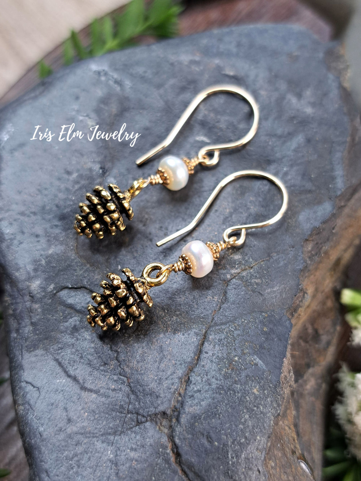A pair of gold-tone dangle earrings with small pinecone charms, white pearl-like beads, and wrapped gold wire details, displayed on a dark textured stone surface. The brand name 'Iris Elm Jewelry' is watermarked on the left side.