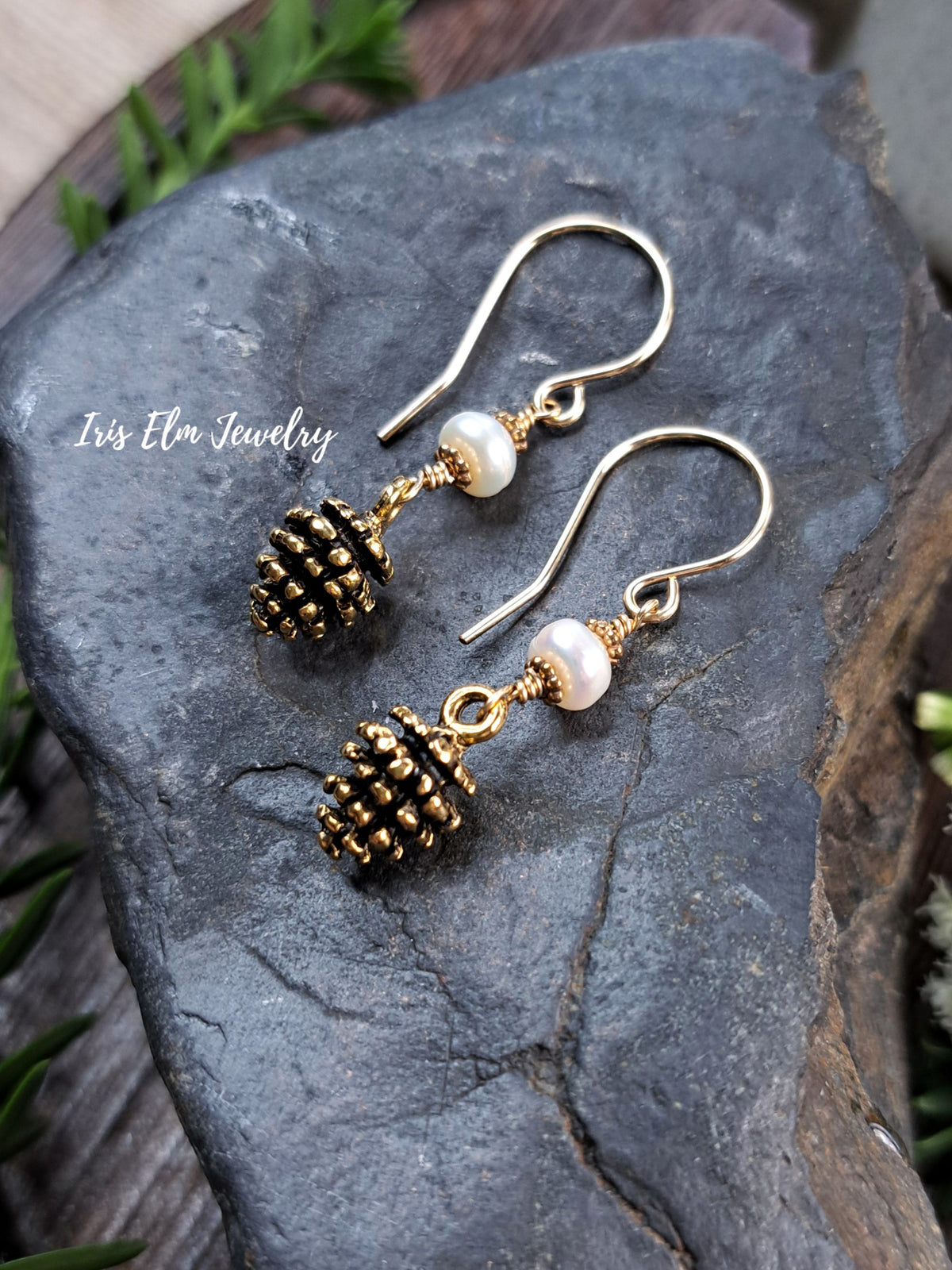 A pair of gold-tone dangle earrings with small pinecone charms, white pearl beads, and curved fishhook wires, displayed on a dark gray rock with green foliage around it. The text 'Iris Elm Jewelry' is visible on the left side.