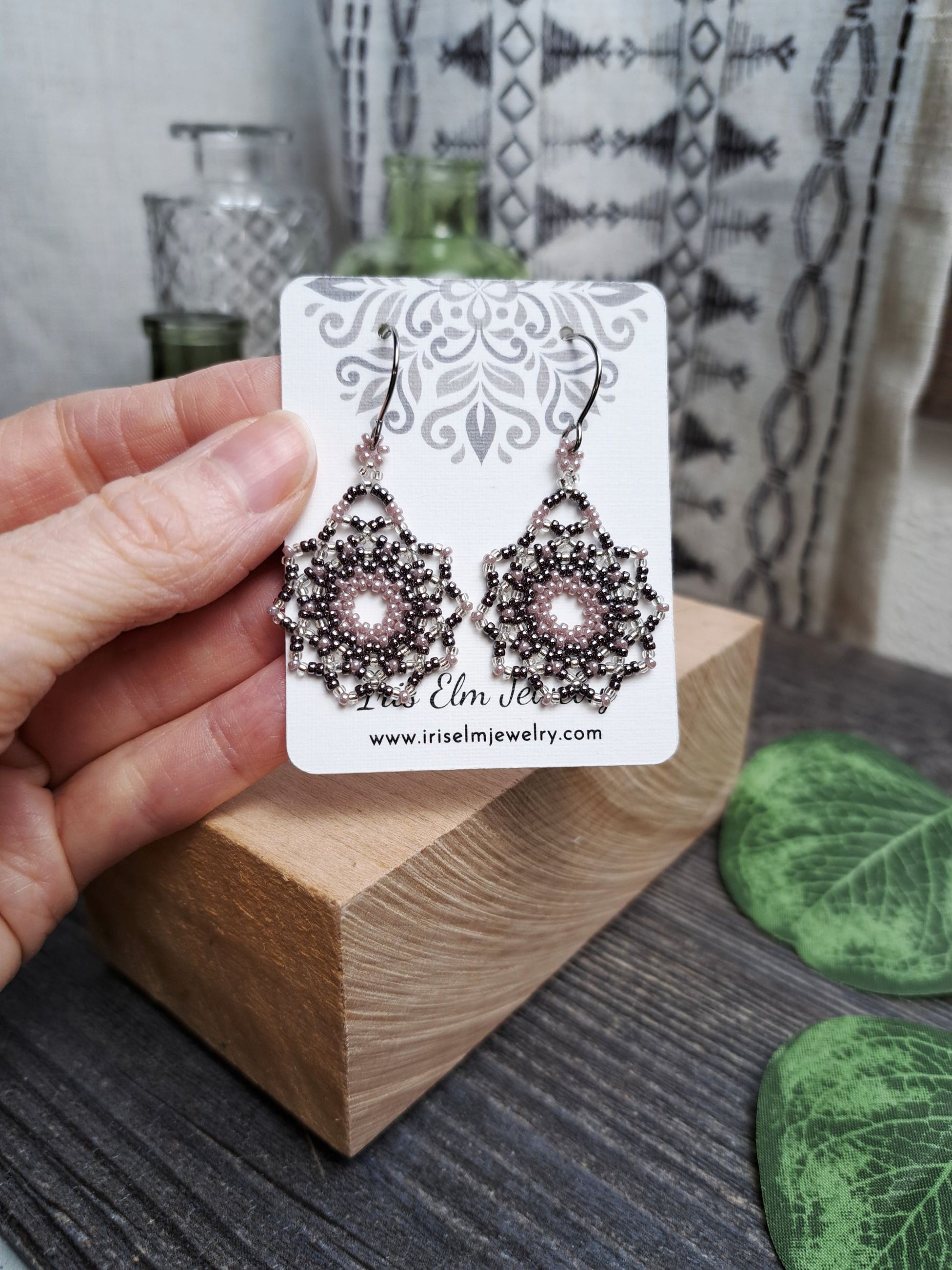 Hand holding a pair of lilac purple seed bead mandala earrings on a card with a wooden block and green leaves in the background