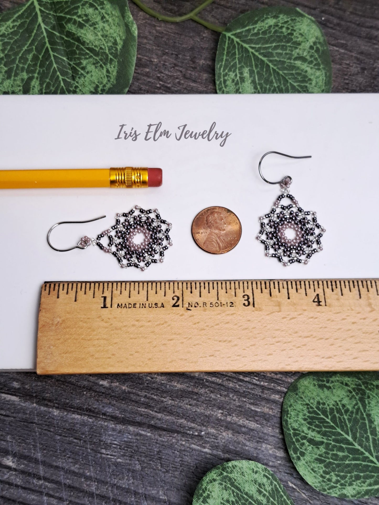 Pair of 1 and 7/8 inch long mandala earrings on a ruler with a penny for scale, on a white card with 'Iris Elm Jewelry' text, surrounded by green leaves.