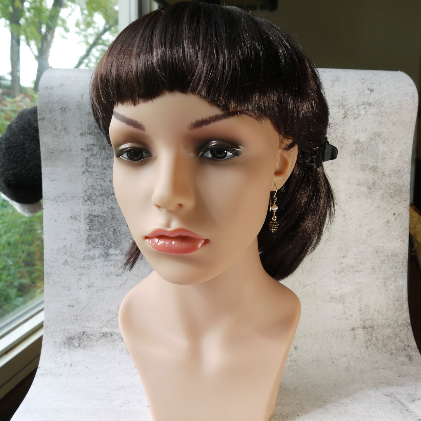 Mannequin head with a dark brown wig wearing pinecone earrings on a textured surface.