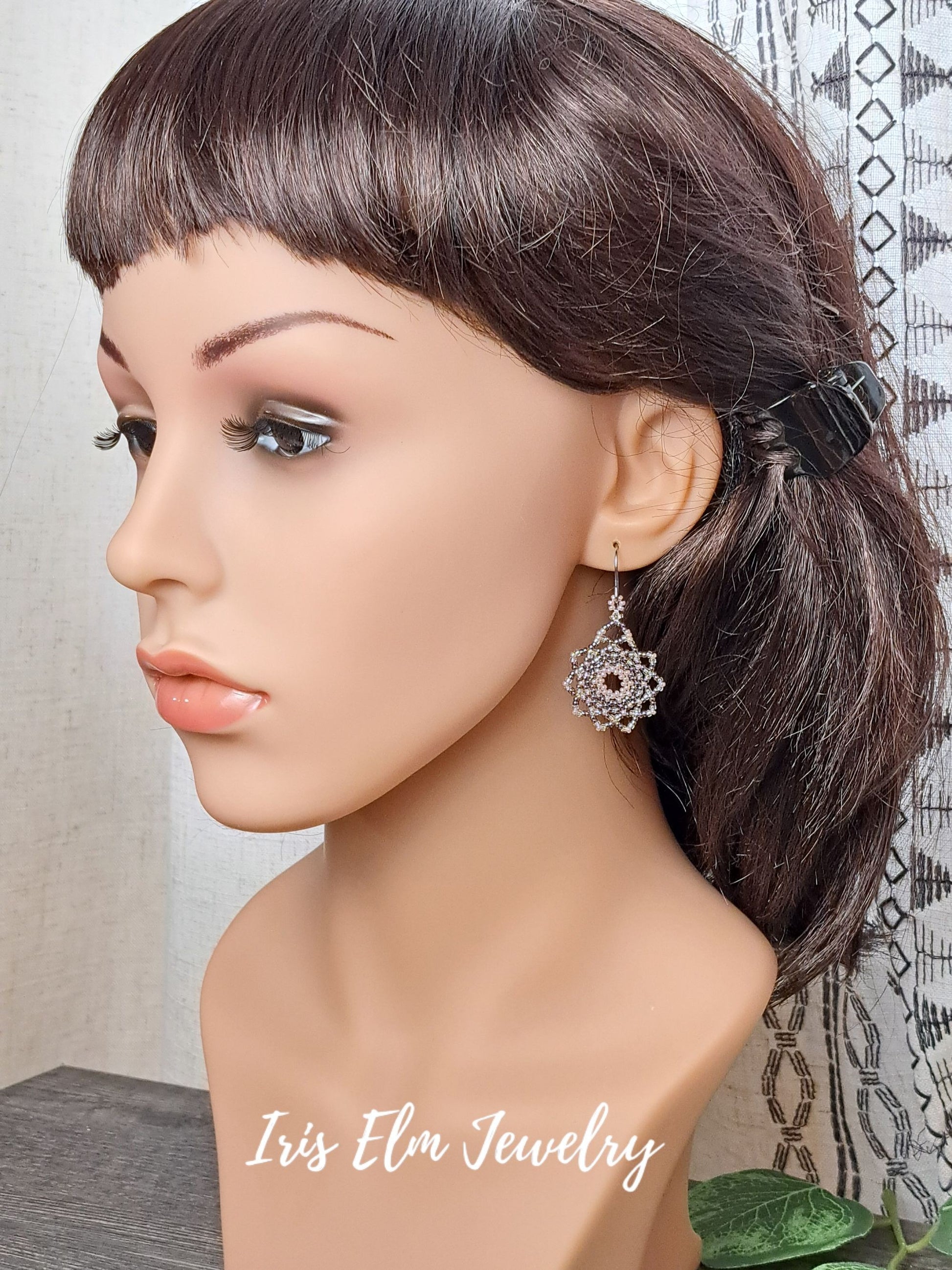 Mannequin head with brown hair styled in a ponytail, wearing a silver lilac purple seed bead mandala earring with a dark stone, against a neutral background.