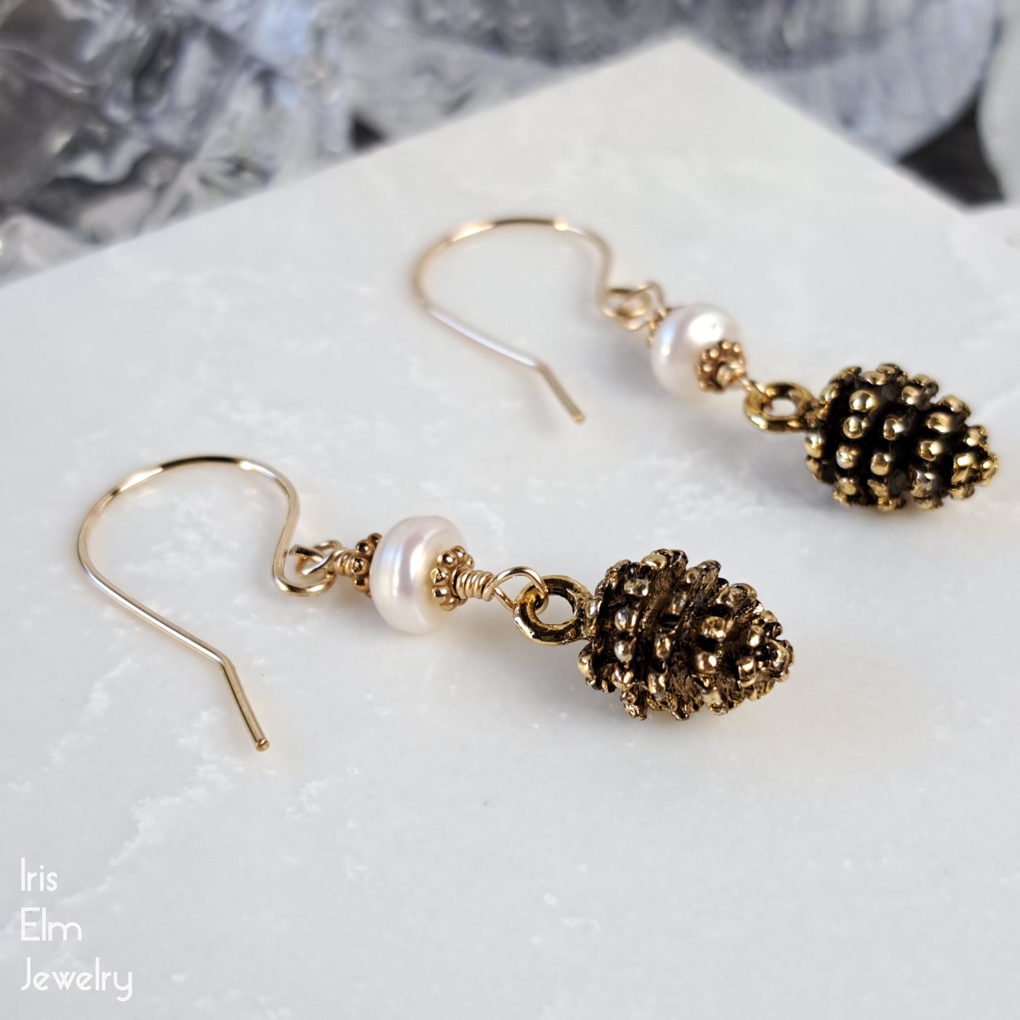 Pair of gold pinecone earrings with pearl accents and goldfilled ear wires on a marble surface, branded 'Iris Elm Jewelry'.