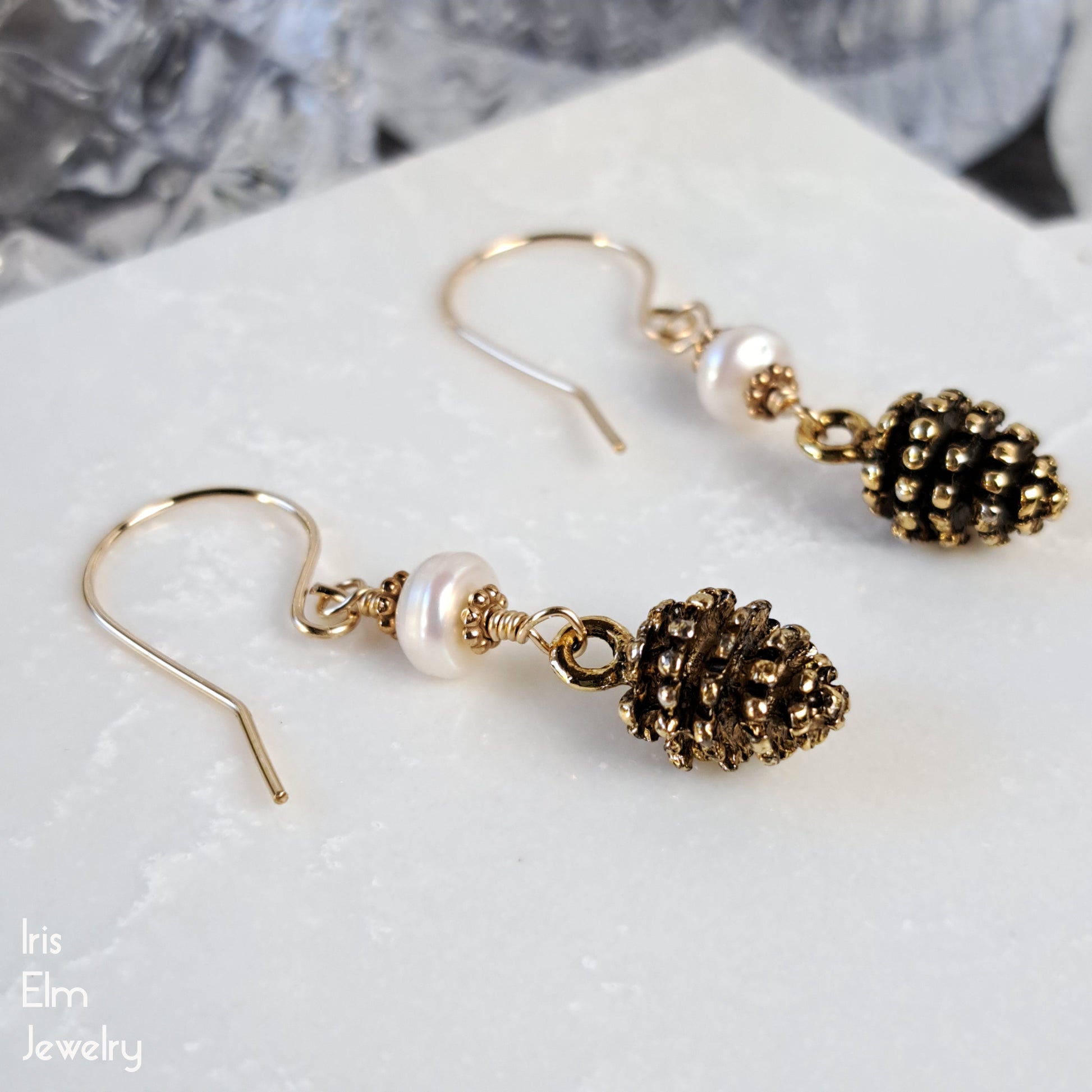 Pair of gold pinecone earrings with pearl accents and goldfilled ear wires on a marble surface, branded 'Iris Elm Jewelry'.