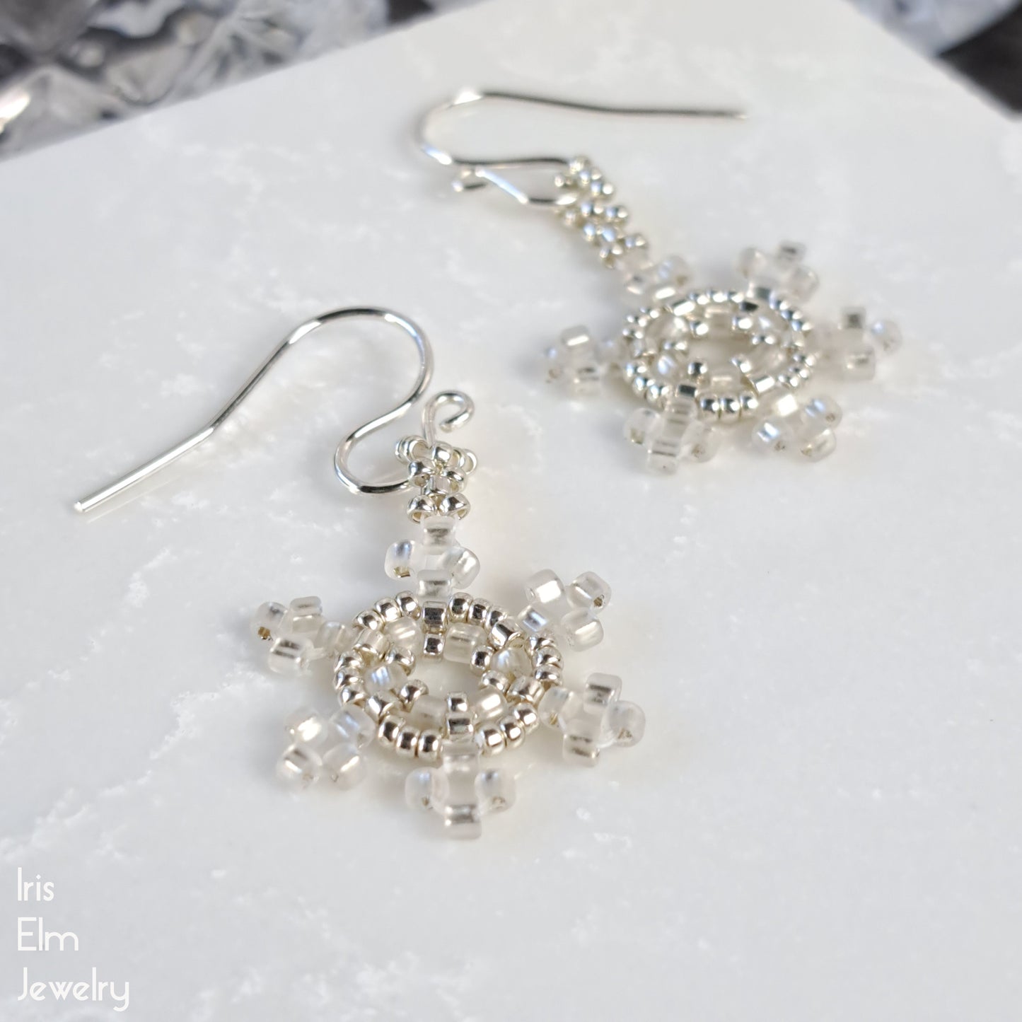 Silver and white snowflake handmade beadwork earrings on a white surface with 'Iris Elm Jewelry' branding.