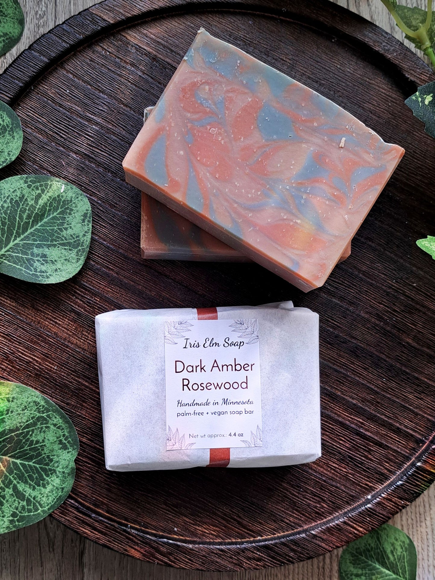 Two dark amber rosewood handmade soap bars with a visible label on a wooden surface surrounded by green leaves.